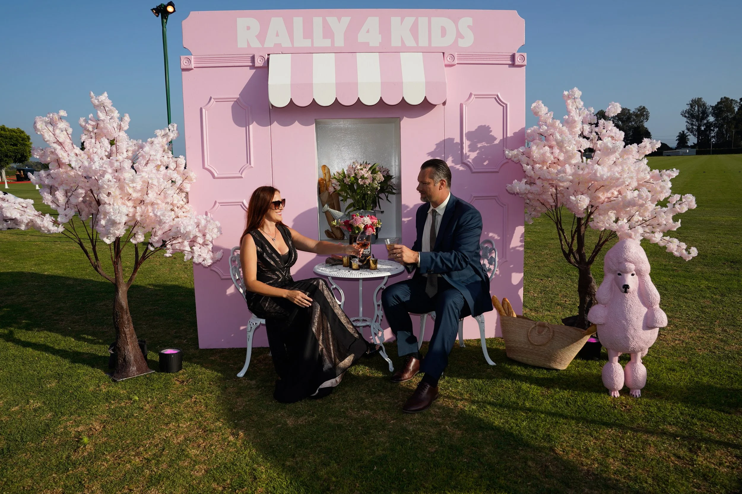 A man and woman sitting at a small round table outside in front of a pink building with the sign 'Rally 4 Kids'. The woman is wearing a black dress and sunglasses, and the man is in a navy suit. They are having drinks and toasting. There are pink flo