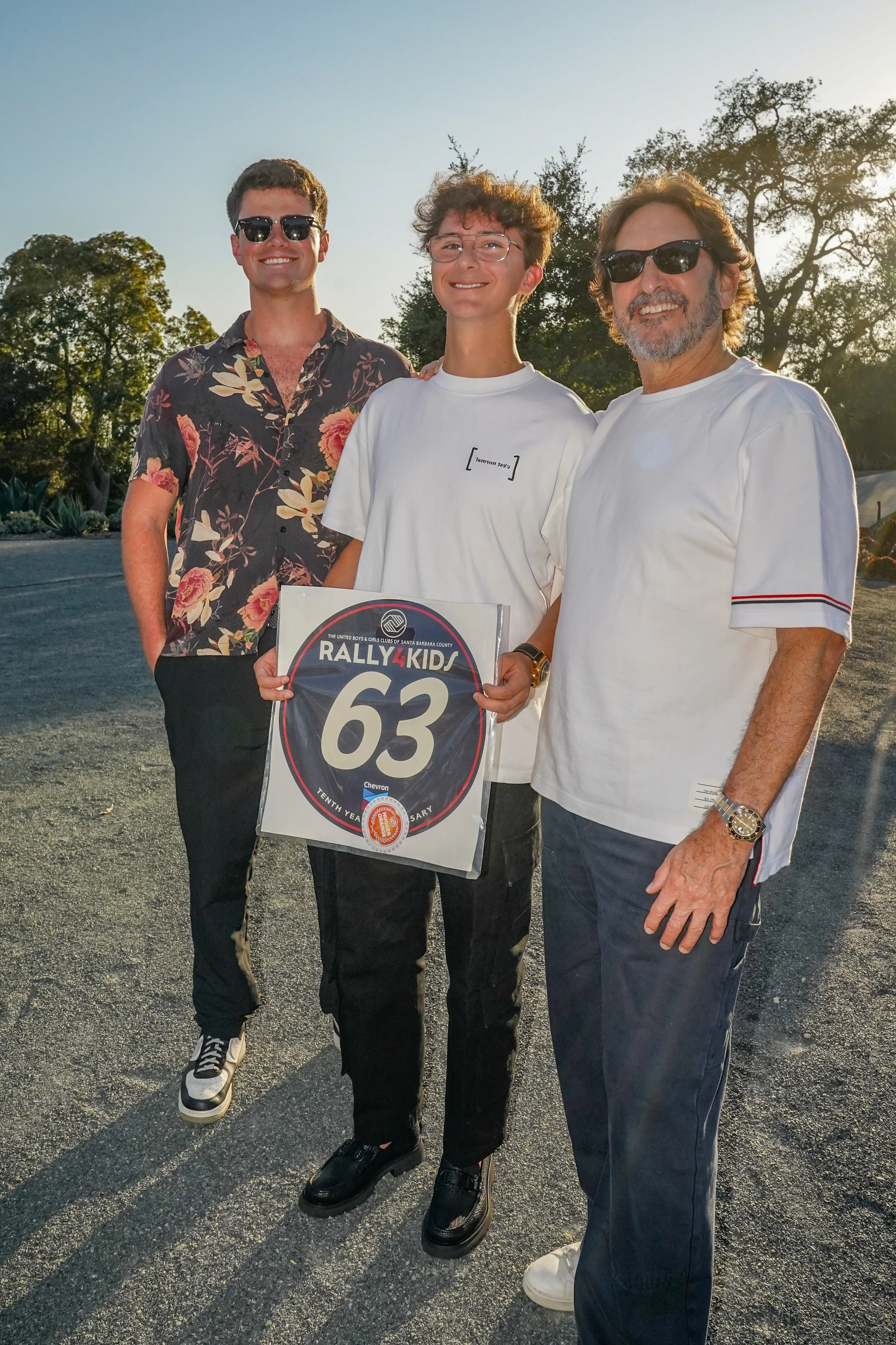 Three males standing outdoors at sunset, one holding a Rally4Kids sign with the number 63. They are smiling and wearing casual clothes, with trees in the background.