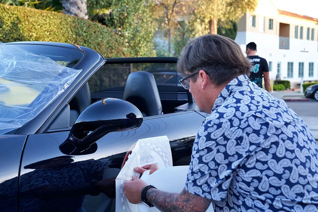 A man with tattoos wearing glasses and a patterned shirt is applying a sticker to a black convertible car. There is a person in a black T-shirt standing in the background near a white building with balconies.