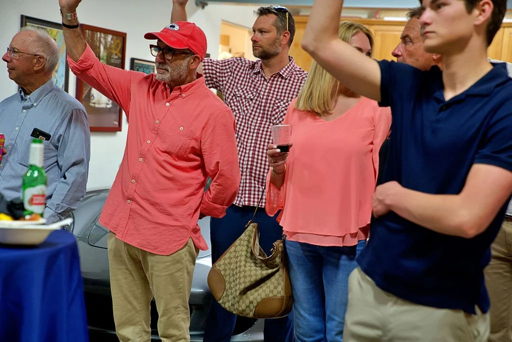 A group of people at a social gathering, one man raising his hand, others holding drinks, indoors with framed pictures on the wall.