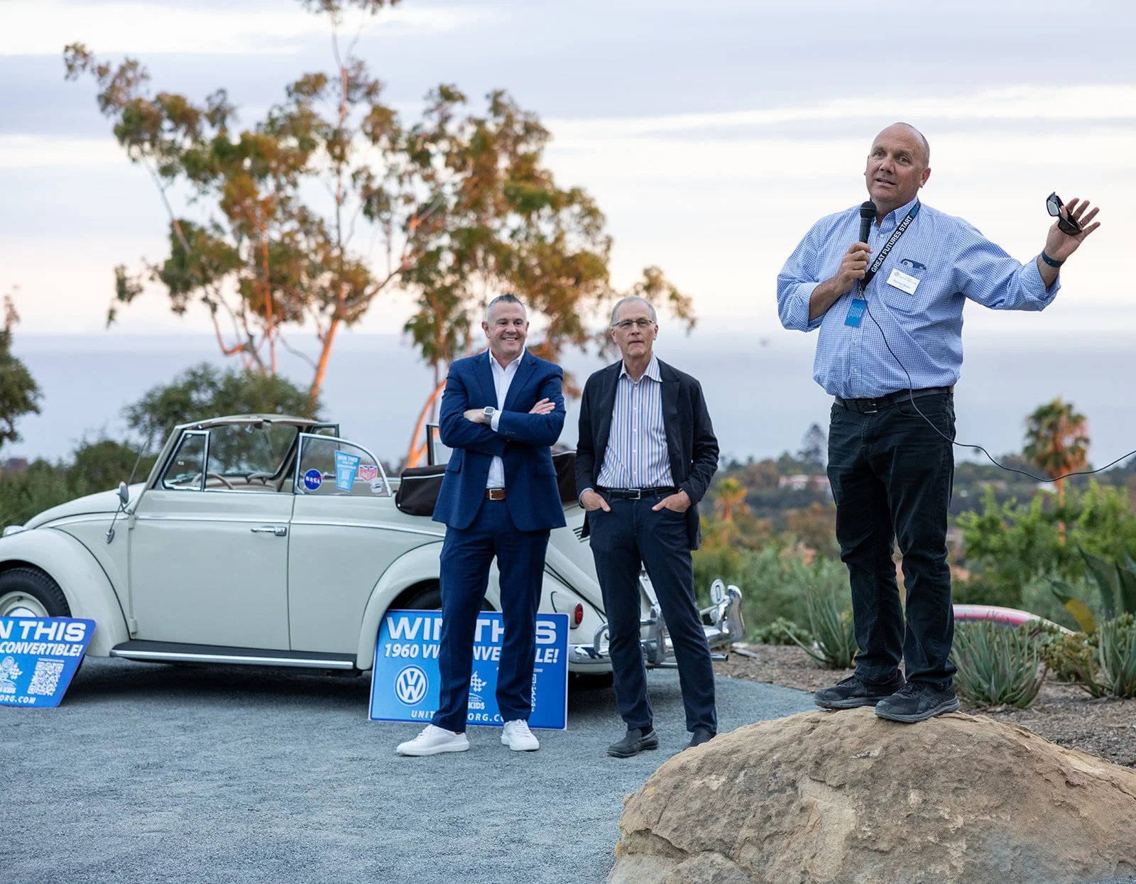 Three men stand outdoors, two wearing suits and one holding a microphone, with a vintage white Volkswagen Beetle behind them and campaign signs that read "Win This 1960 VW Convertible!" in the foreground. The scene includes trees and a cloudy sky in 