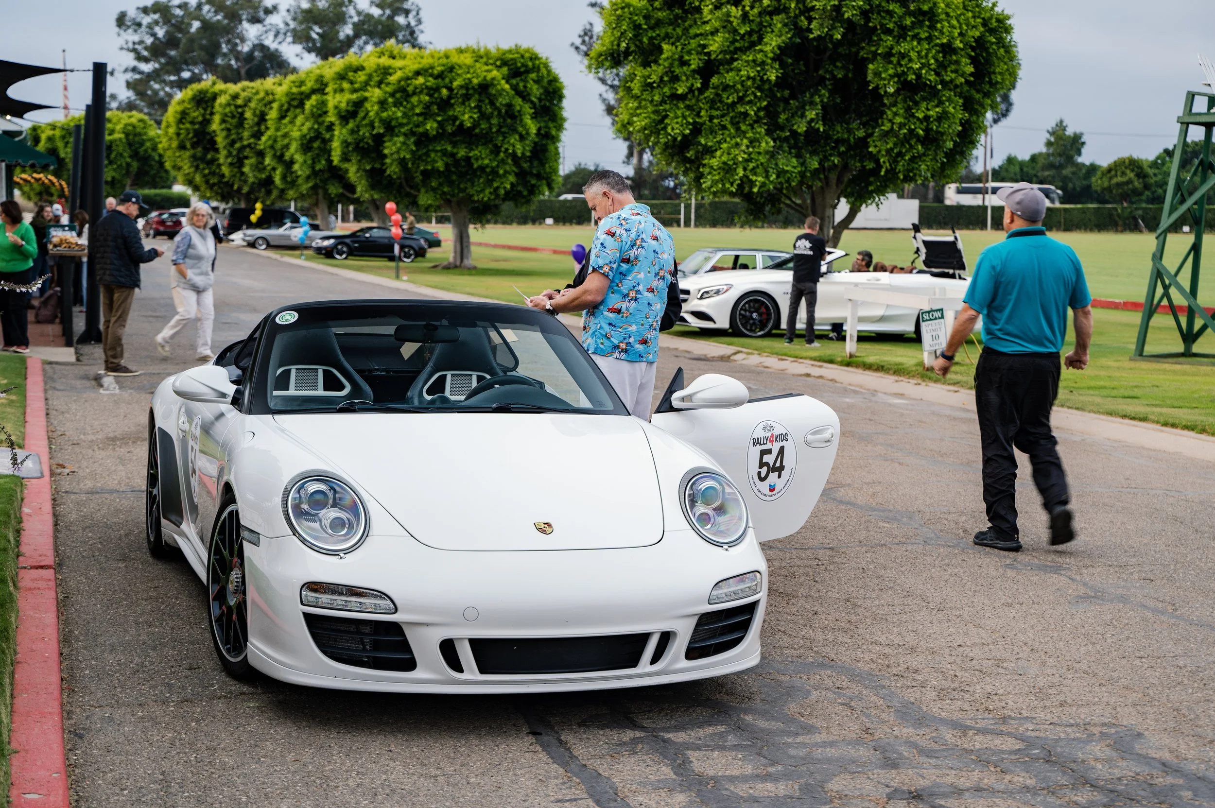 A white Porsche sports car with a black convertible top, displayed at an outdoor event. The car has a sticker on the front that says "Rally 4 Kids" and the number 54. People are walking around, with some looking at the car and others in the backgroun