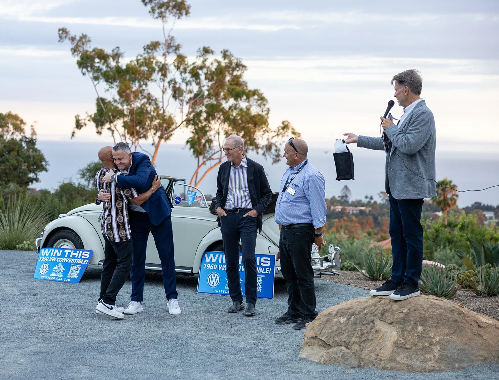 Group of men at an outdoor event with a vintage Volkswagen convertible car and signs promoting a contest to win the car. One man is holding a black gift bag, one is using a microphone, and two others are hugging and standing near the car. Trees and a