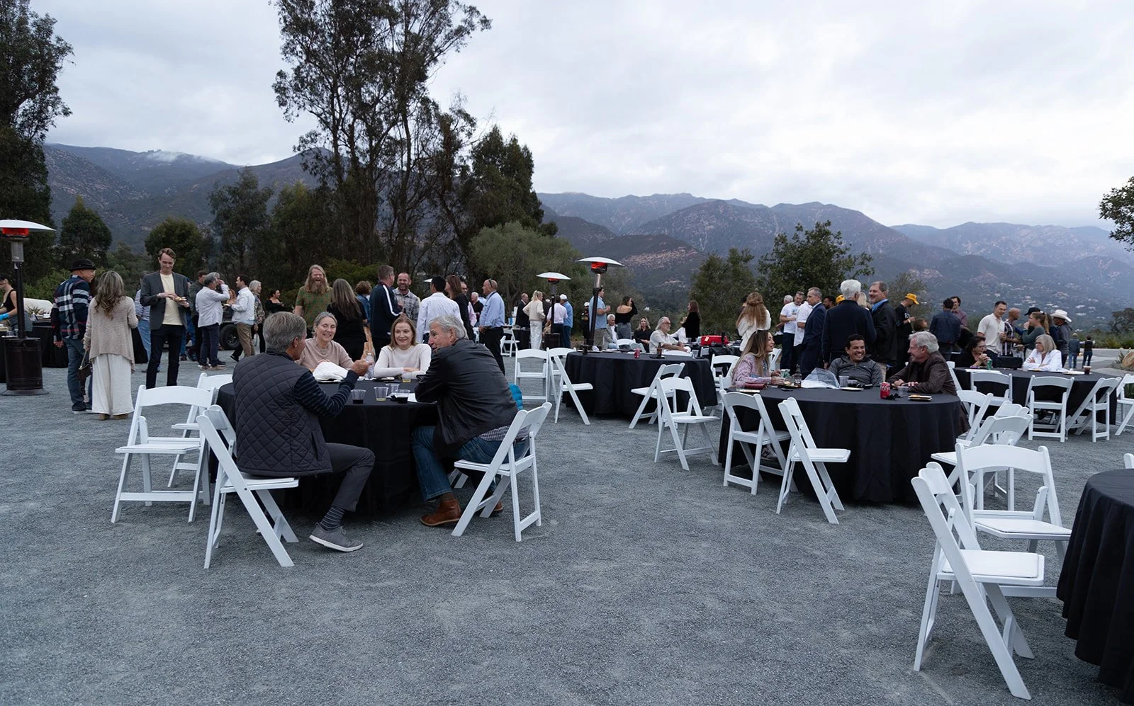 People gathering at an outdoor event with tables and chairs, mountains in the background, and cloudy sky overhead.