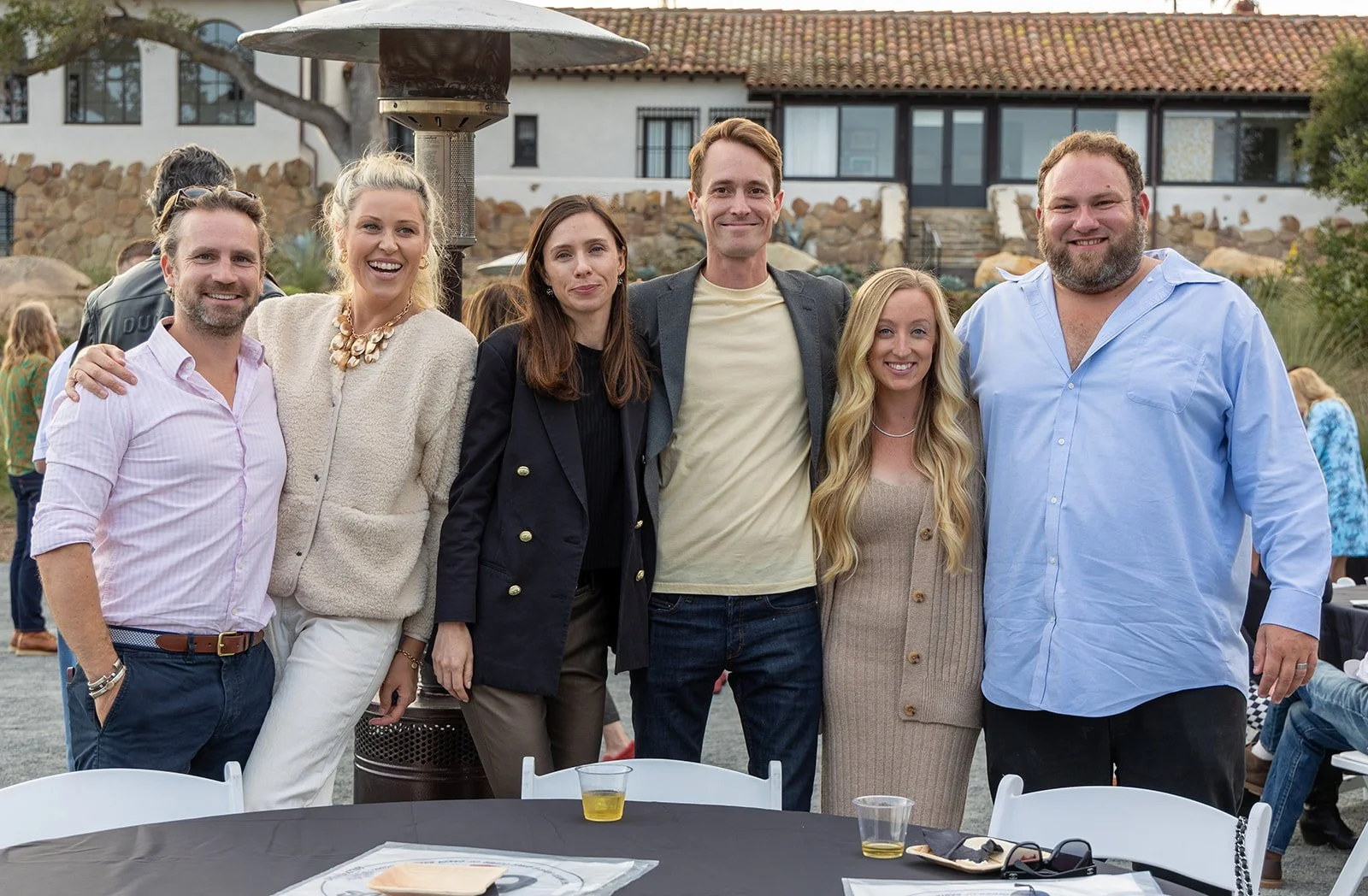Group of six people smiling and posing outdoors at a social event, with a building and trees in the background.