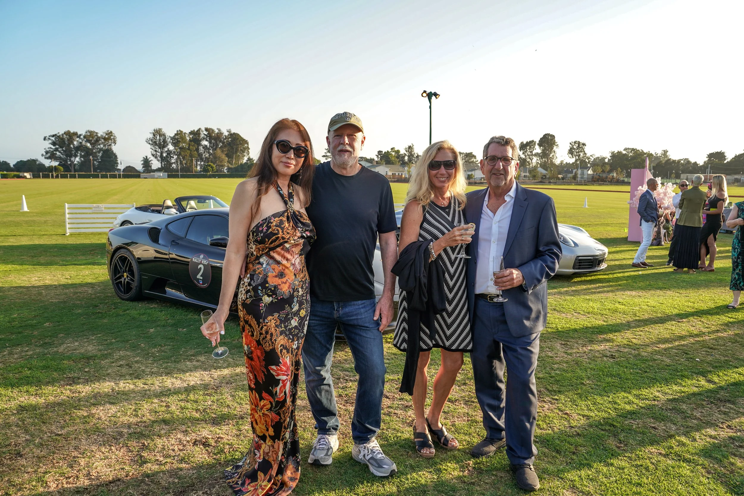 Four people standing on a grassy field at a car event during sunset. Two women and two men, all dressed in semi-formal attire and holding glasses. Classic and sports cars in the background.