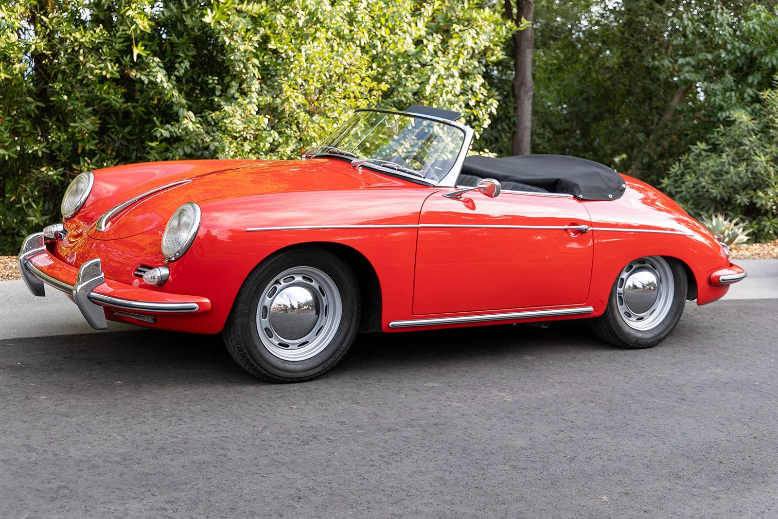 Red vintage Porsche 356 convertible car parked on a paved driveway with green trees and bushes in the background.