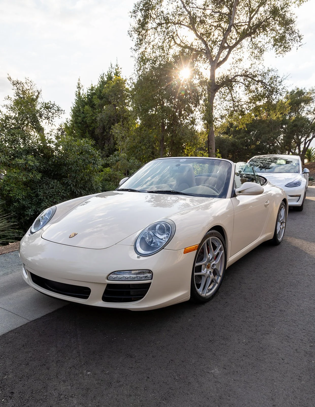 A white Porsche convertible parked on a street with trees and the sun shining behind them.