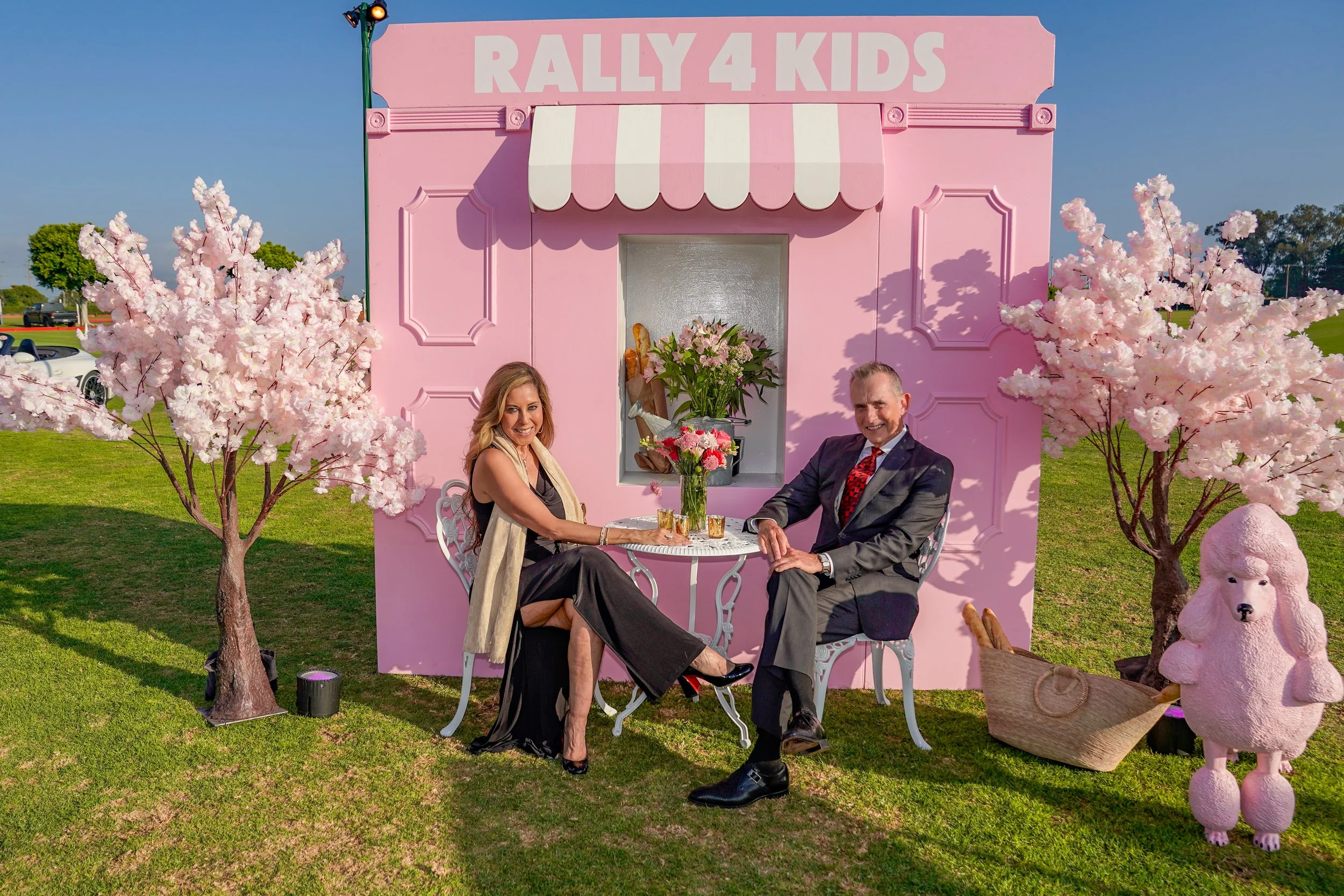 A man and woman sitting at a small white table outdoors in front of a pink building with the sign "Rally 4 Kids." The woman is wearing a black dress and scarf, and the man is in a suit and tie. They are smiling and holding hands. There are pink flowe