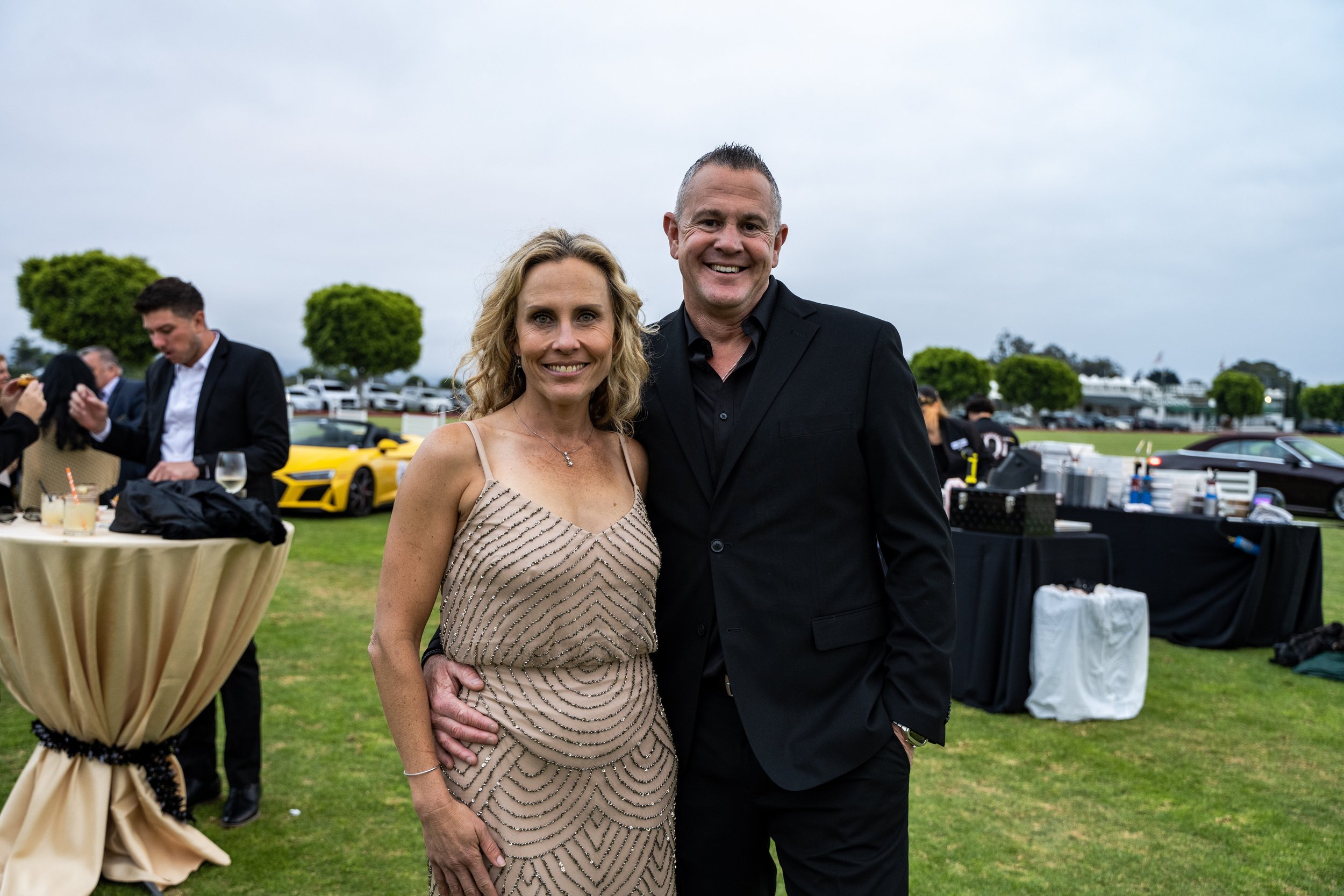 A woman in a beige dress with a beaded pattern and a man in a black suit standing together outdoors at a formal event, with cars, trees, and guests in the background.
