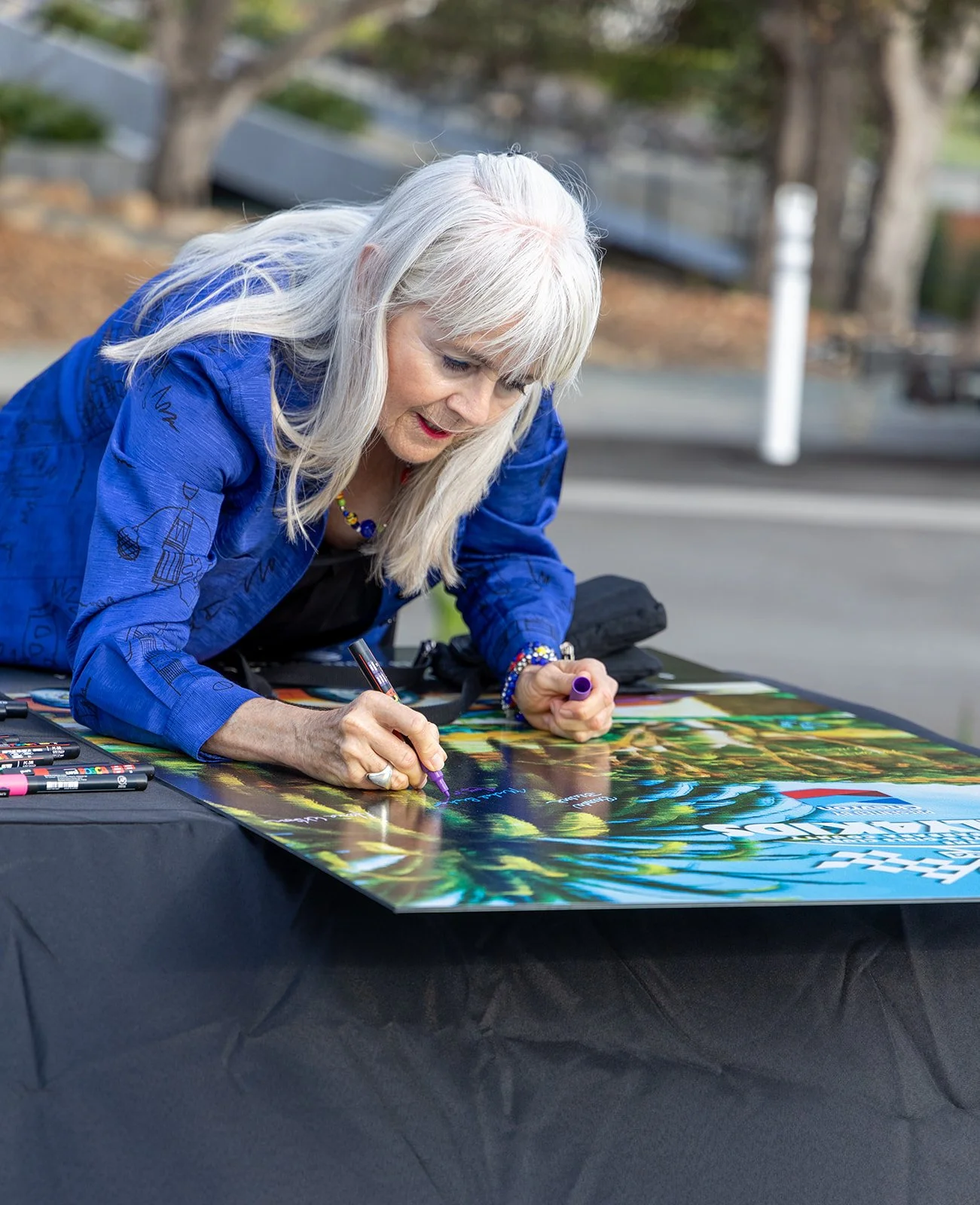 An older woman with long gray hair, wearing a blue jacket, is signing a colorful poster on a black table outdoors.