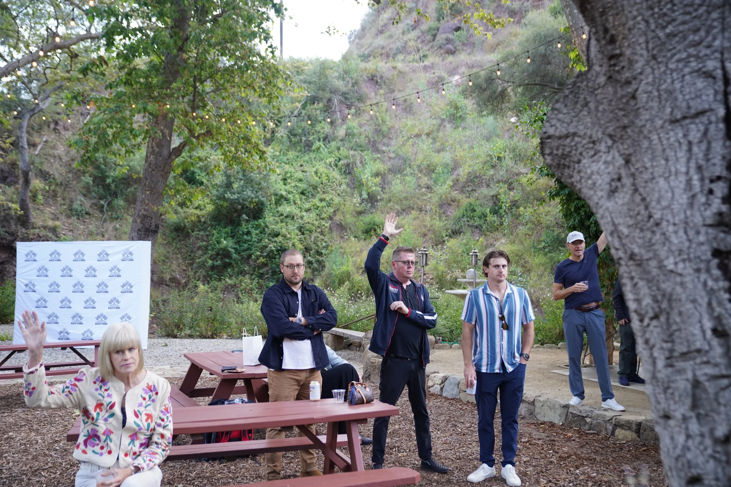 A group of six people at an outdoor event in a wooded area, with tiered seating, a backdrop, and string lights overhead. One woman is sitting at a table with her hand raised, while four men stand, one raising his hand, and another holding a drink.