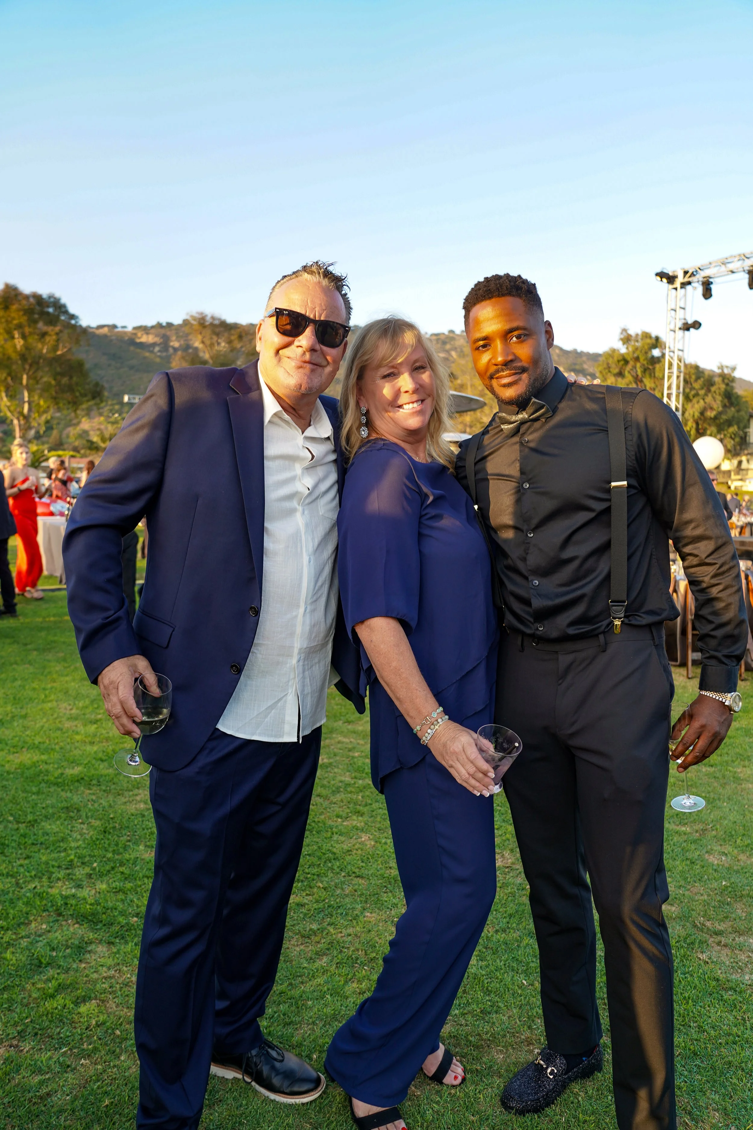 Three people posing outdoors at an event during the daytime. They are dressed in formal attire, holding drinks, and smiling at the camera. The background features other guests, tables, and a scenic hillside with trees.