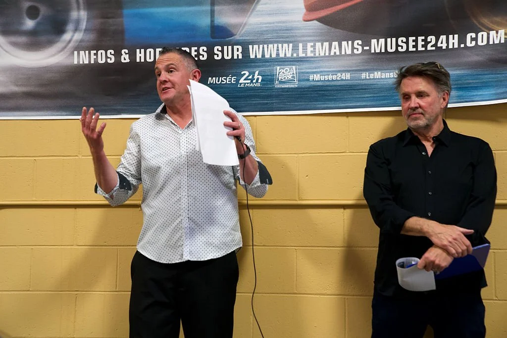 Two men standing in front of a banner at an event. The man on the left is speaking and holding papers, while the man on the right is listening with his arms crossed. The banner promotes the Le Mans Museum website.