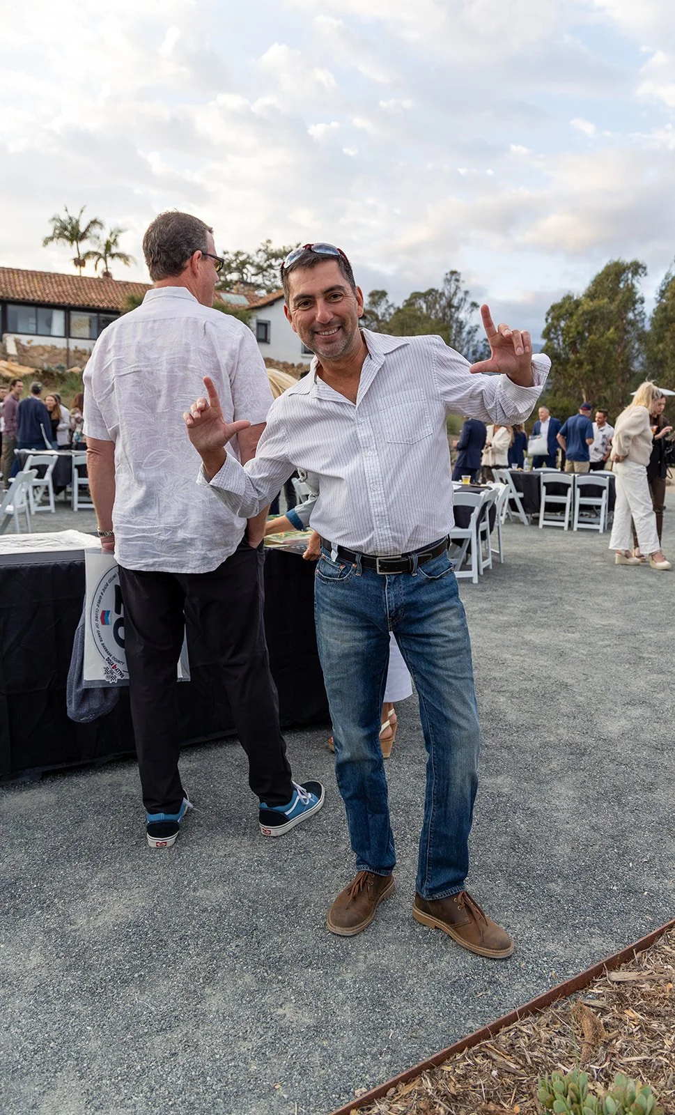 A man in a white striped shirt and jeans smiling and making a finger gun gesture at an outdoor event with other people in the background.