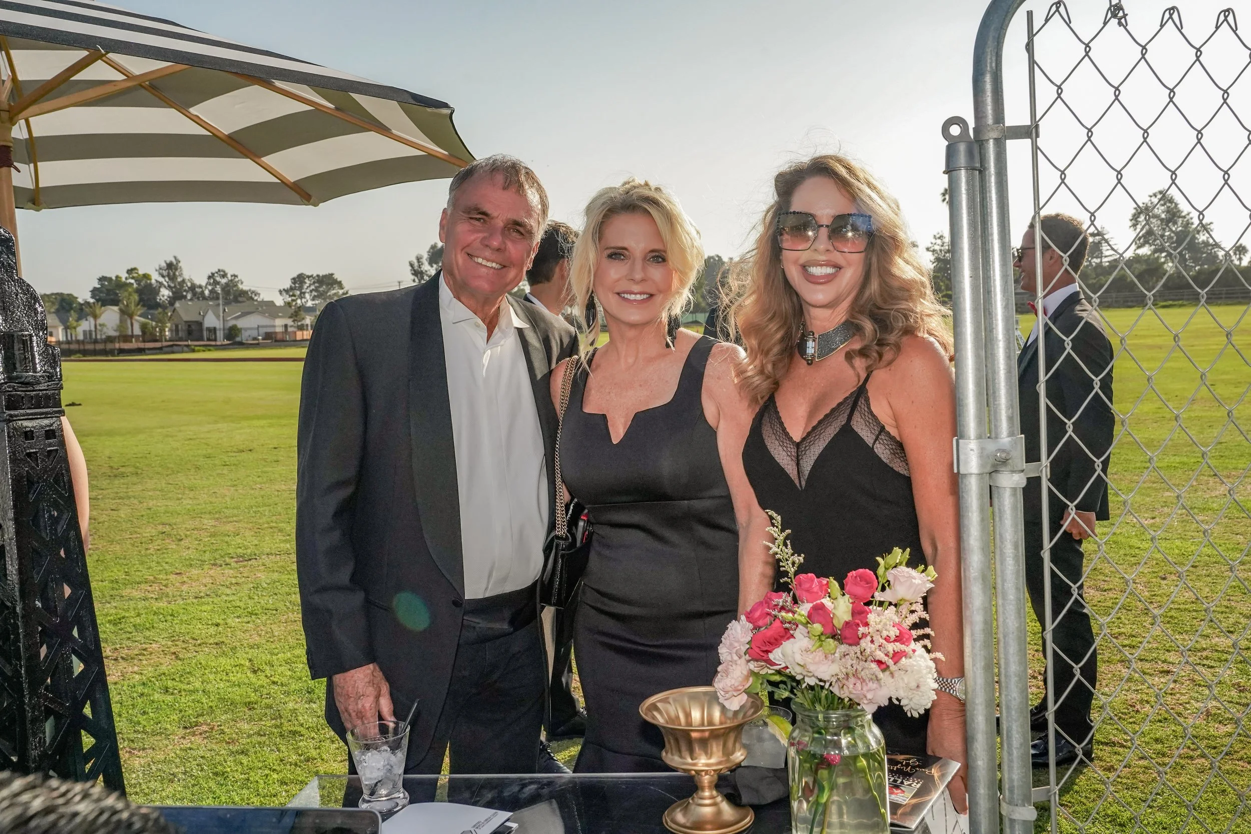 Three people, two women and one man, dressed in formal attire, posing together outdoors on a sunny day, with a grassy field and buildings in the background. The woman on the right is holding a bouquet of pink and white flowers.