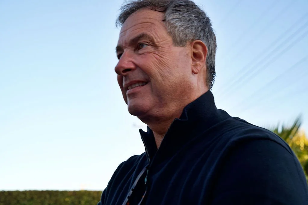 A middle-aged man with short gray hair smiling outdoors on a clear day, wearing a black jacket.