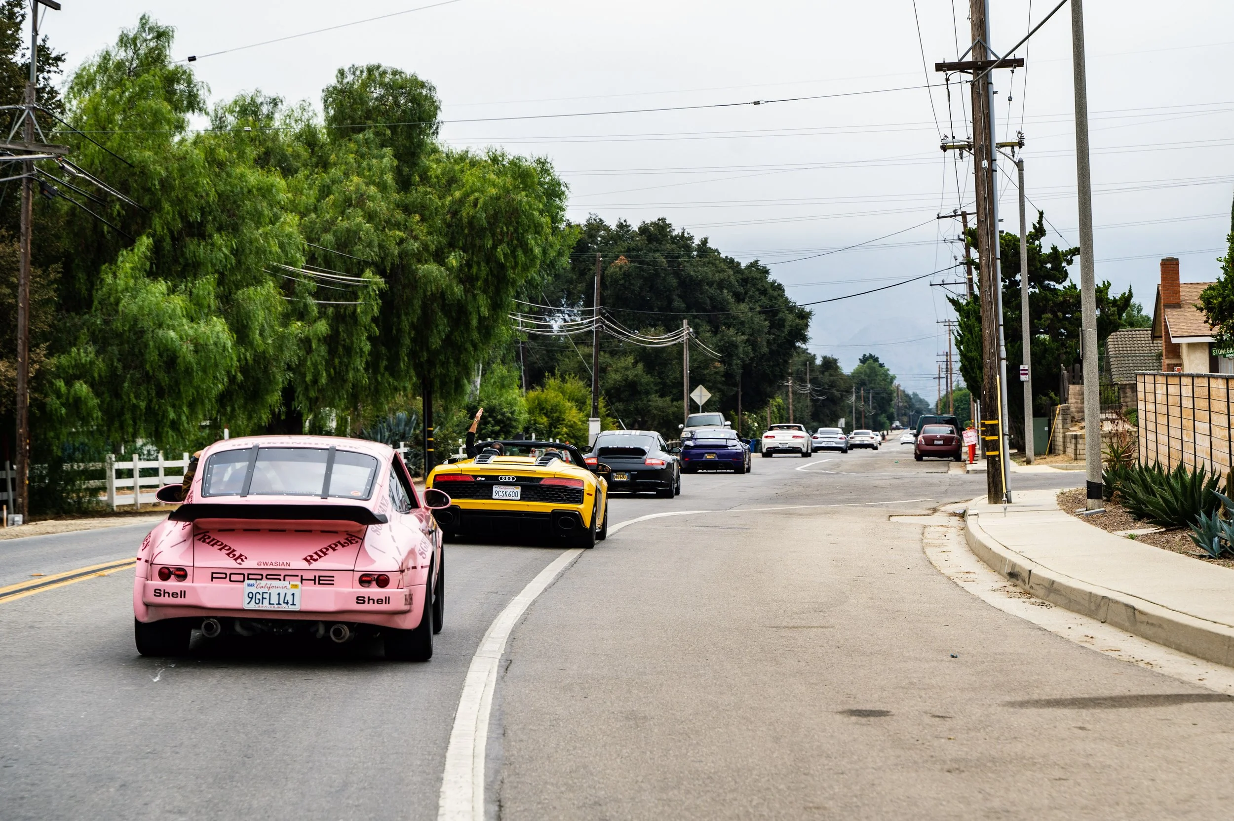 Multiple cars, including a pink Porsche and a yellow Audi, driving on a street with trees and utility poles along the side.