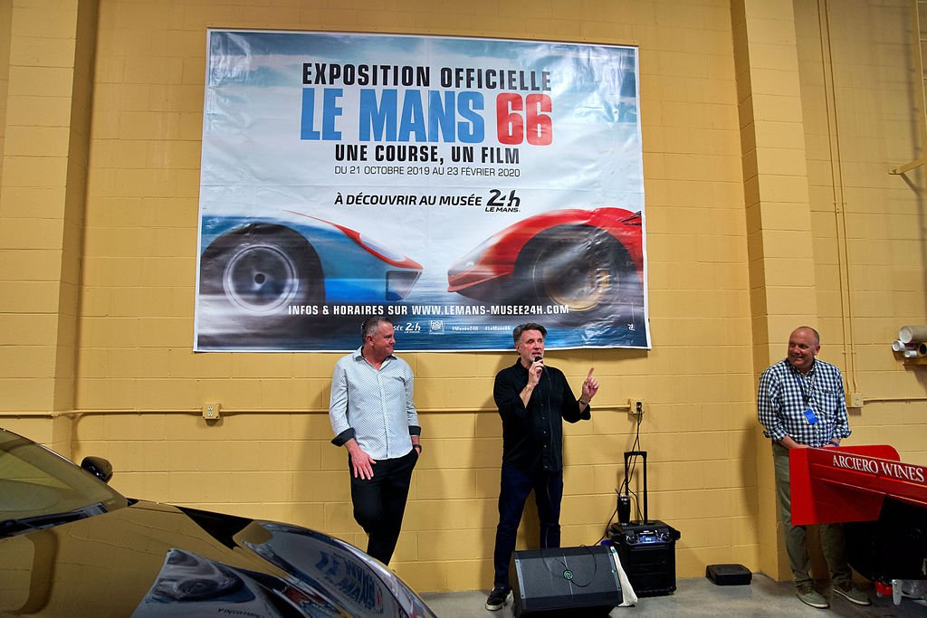 Three men standing in front of a large poster about the Le Mans 66 exhibition, with one man speaking into a microphone. A golden car is partially visible in the foreground, and there is a red Argiero Wines display on the right.