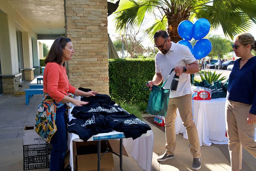 People shopping at an outdoor table with black T-shirts, under a palm tree, with balloons and gift bags nearby.