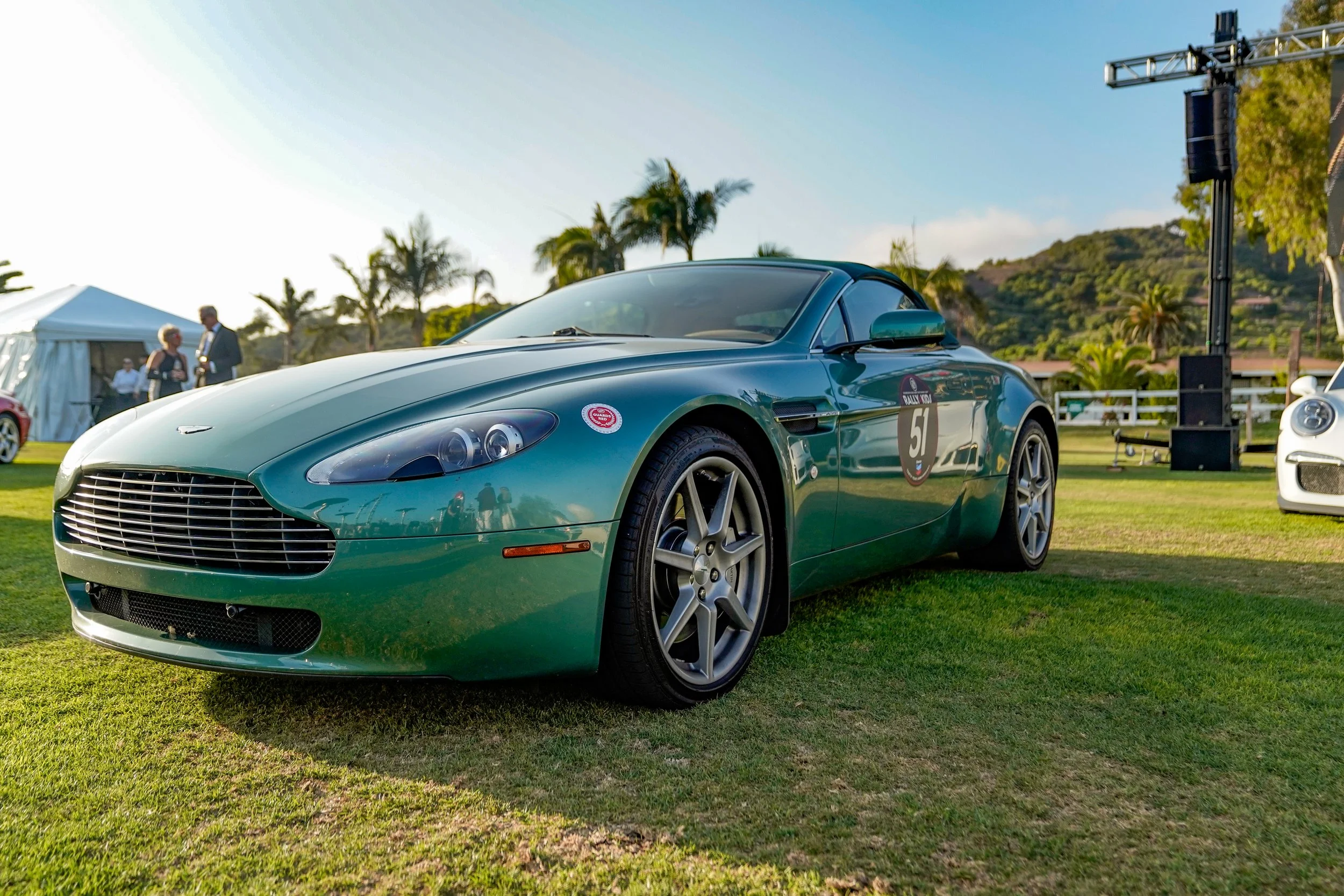 A classic green Aston Martin sports car displayed outdoors at a car event, with people, tents, trees, and a mountain in the background.