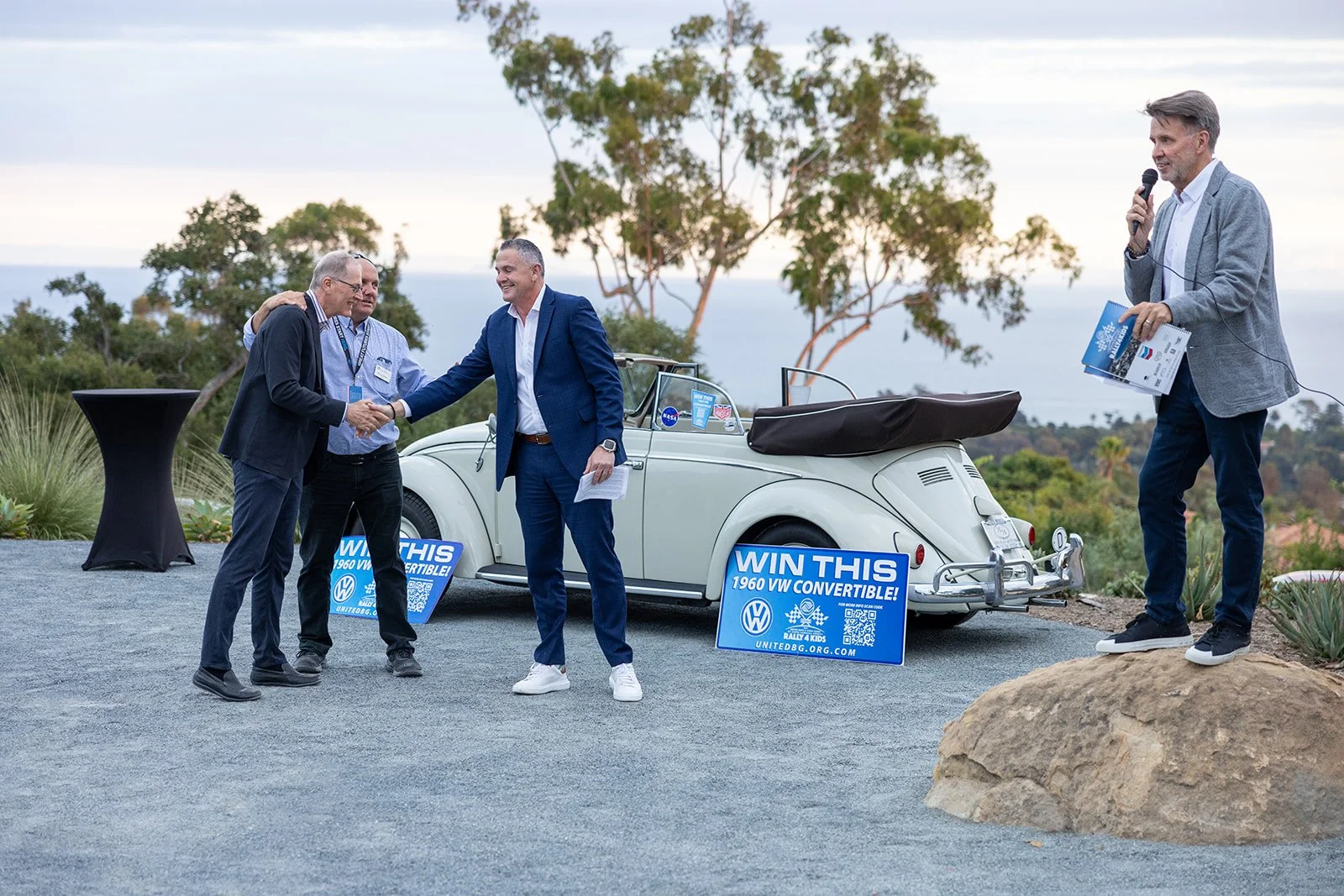 Four men at an outdoor event, two shaking hands, with a vintage white Volkswagen convertible car behind them. Signs reading 'WIN THIS 1960 VW CONVERTIBLE!' are placed on the ground. One man is speaking into a microphone.