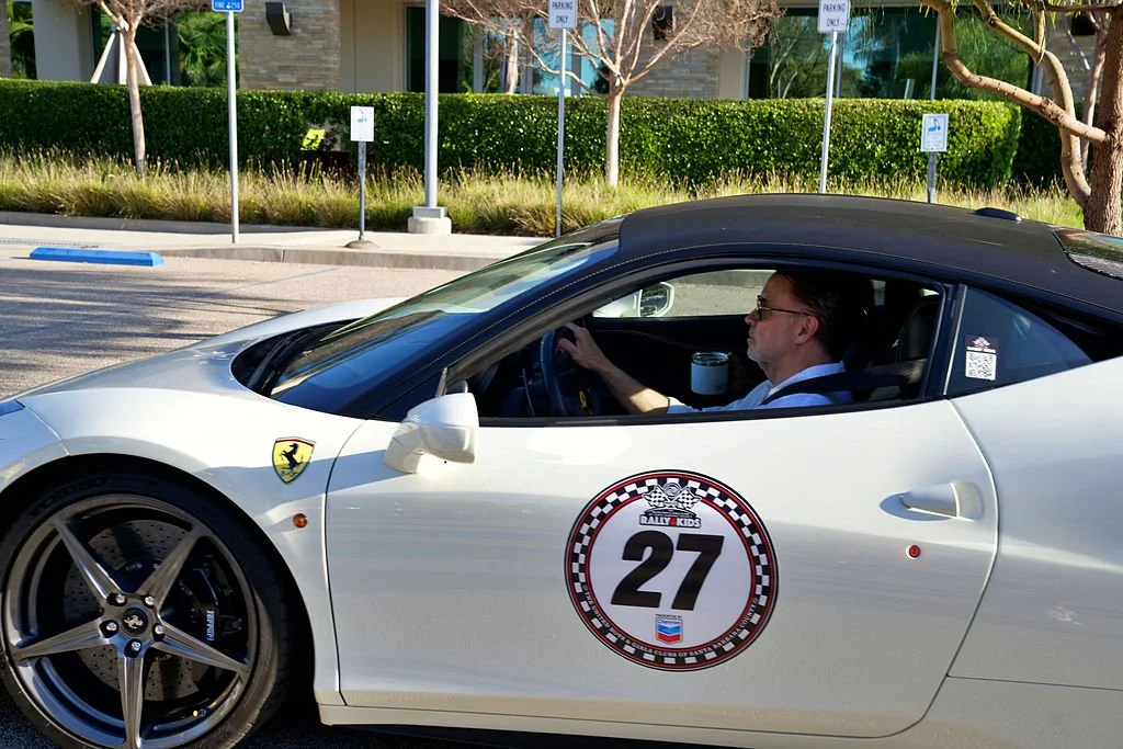 A man driving a white Ferrari with a black roof, displaying a race number 27 and a Rally Kids logo on the door, is in a parking lot surrounded by trees and signs.