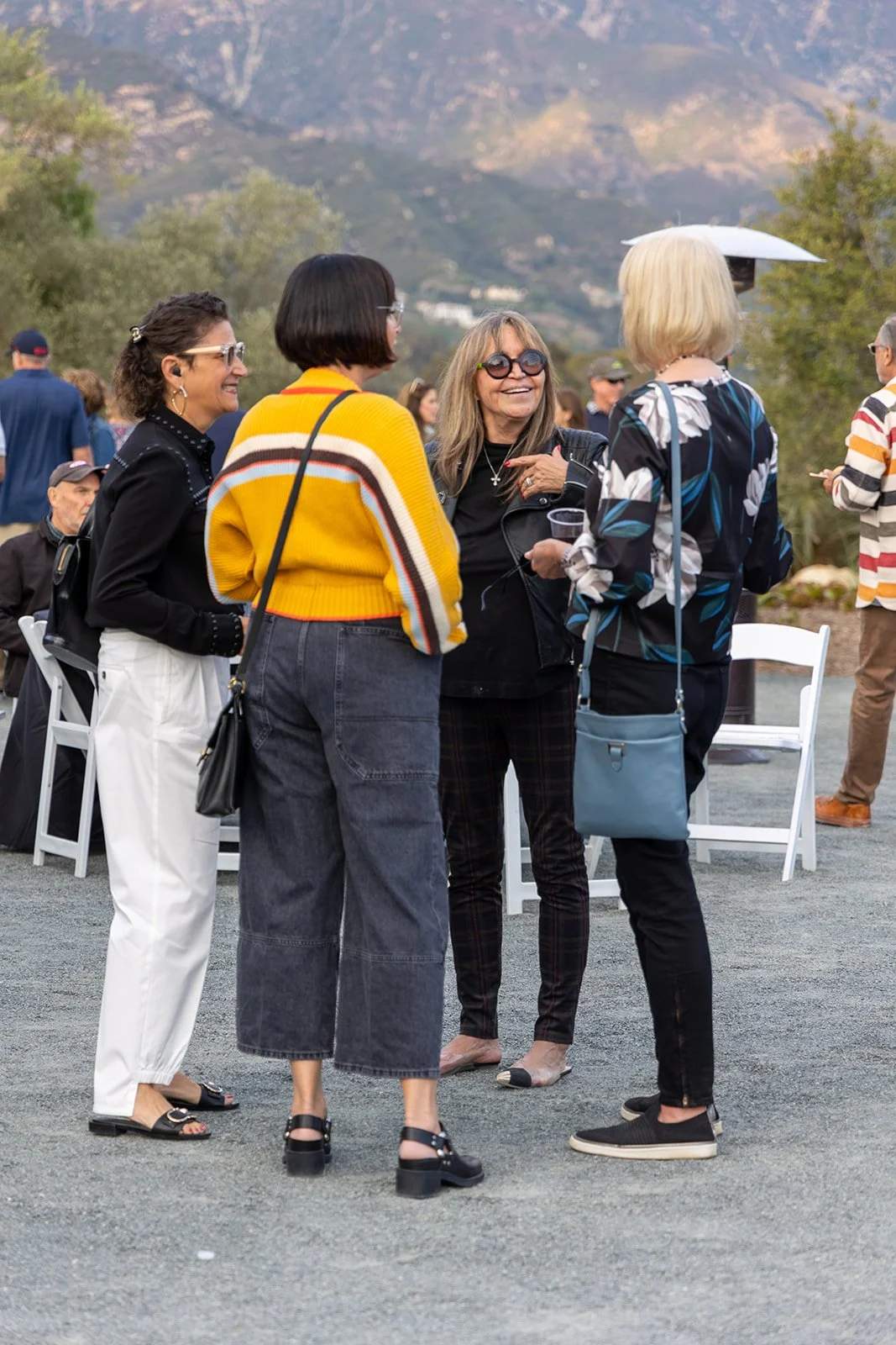 Four women are standing and talking outdoors at a social event with mountains in the background. They are dressed casually and smiling, with some wearing sunglasses and carrying bags.