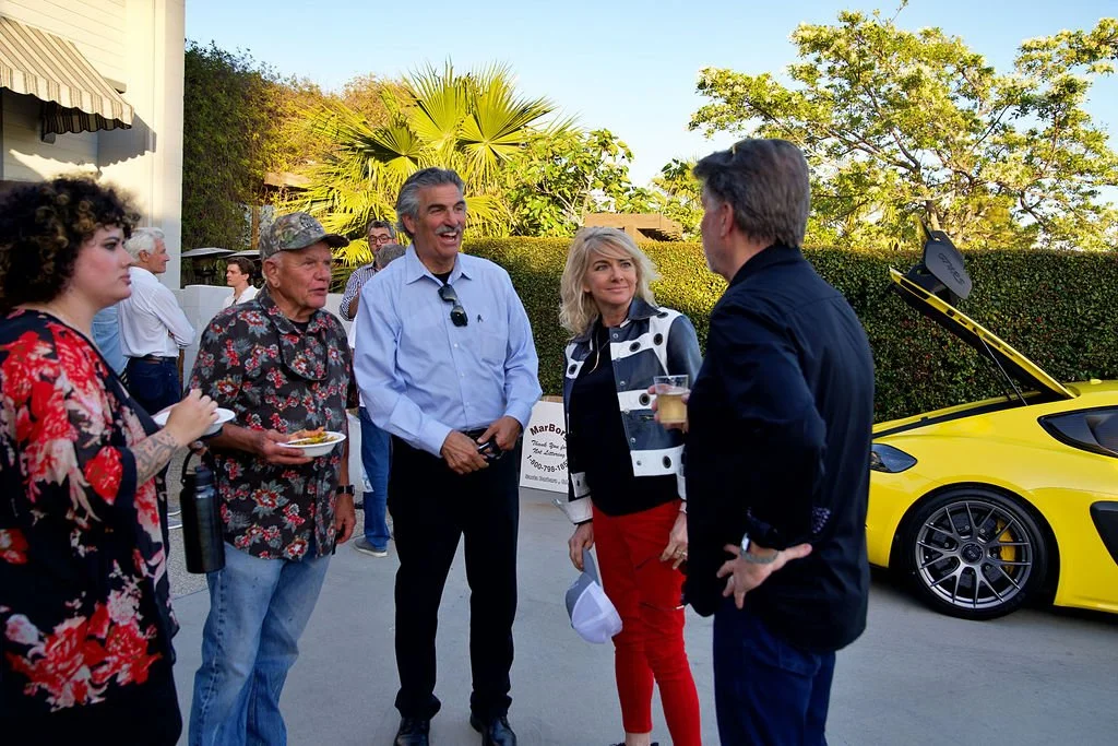 Group of five people having a conversation outdoors next to a yellow sports car with its trunk open. The background includes trees and a building.