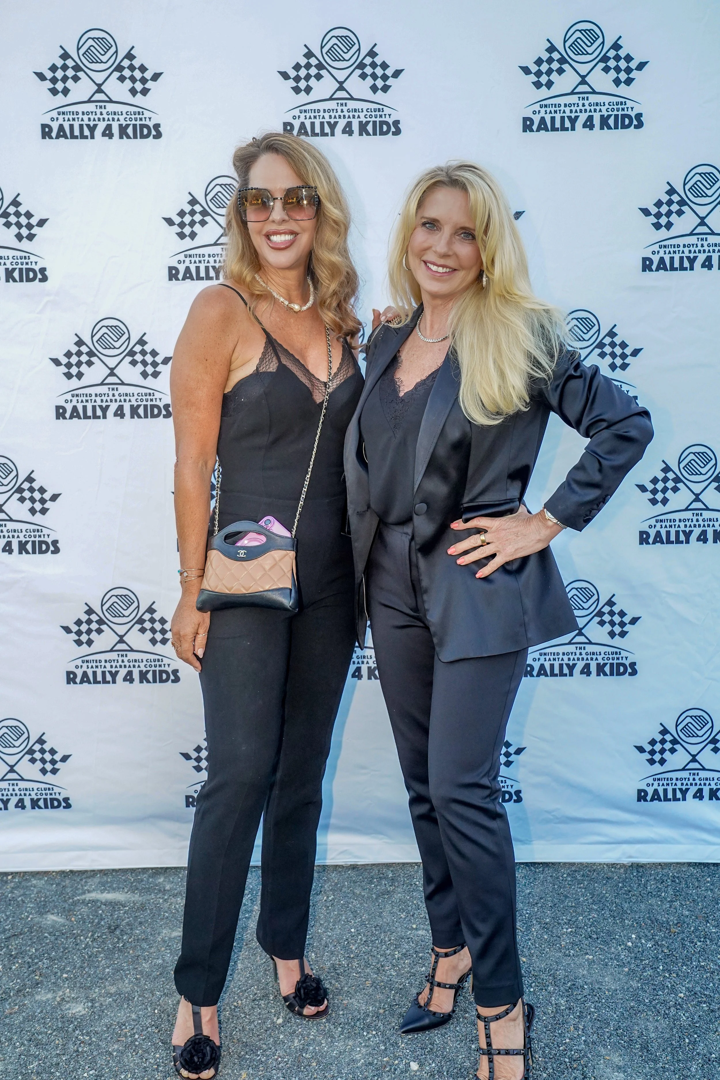 Two women standing in front of a backdrop with a racing-themed logo that reads "The United Boys & Girls Clubs of Santa Barbara County Rally 4 Kids." They are dressed in black attire, smiling, and posing for the camera.