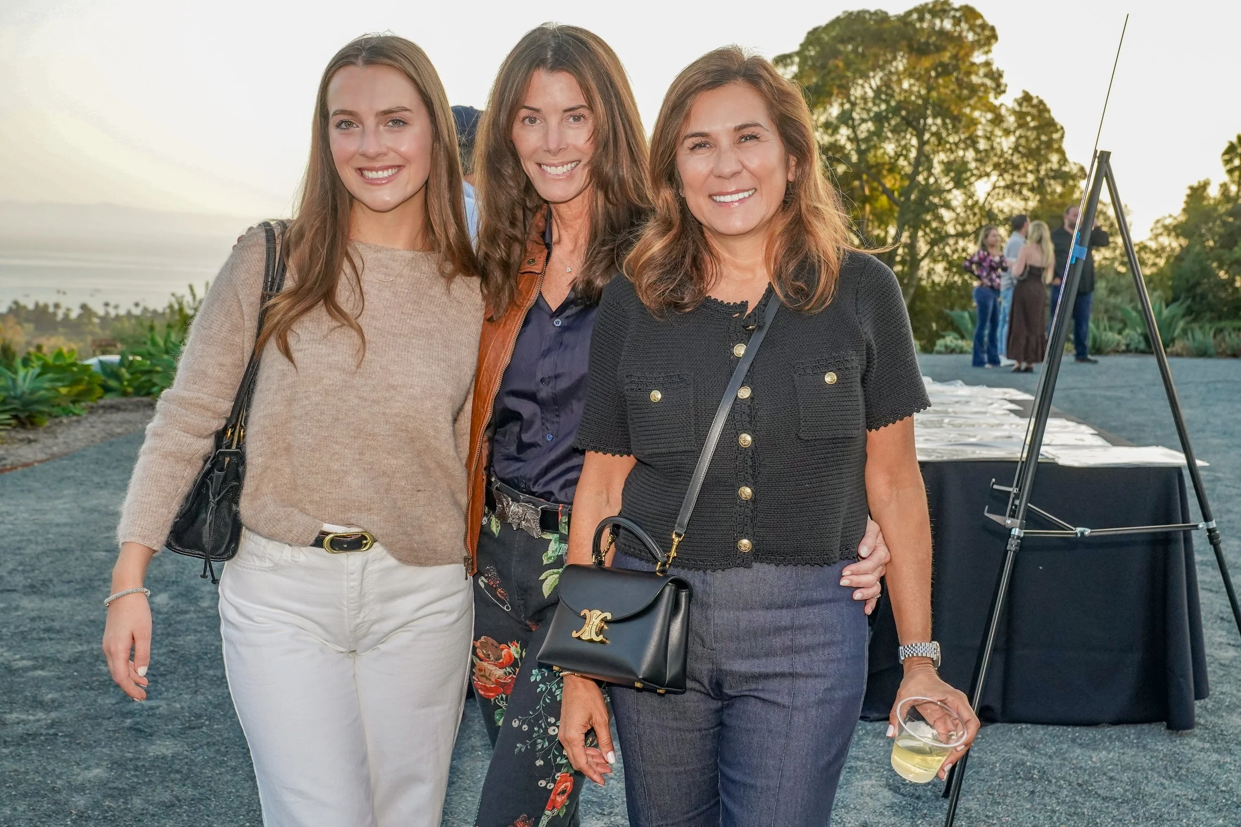 Three women smiling at an outdoor event during sunset, with a scenic landscape and other groups of people in the background.