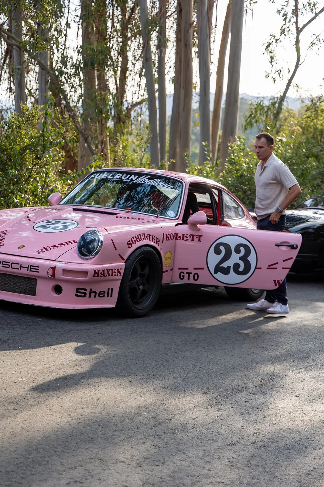 A man standing next to a pink Porsche race car with the number 23, surrounded by trees.