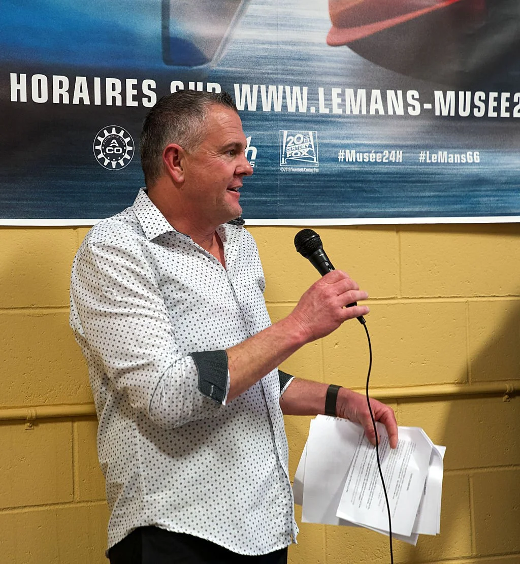 A man with short gray hair, wearing a white polka dot shirt, is speaking into a microphone while holding papers in his left hand, standing in front of a yellow wall and a large blue banner with website information and hashtags.