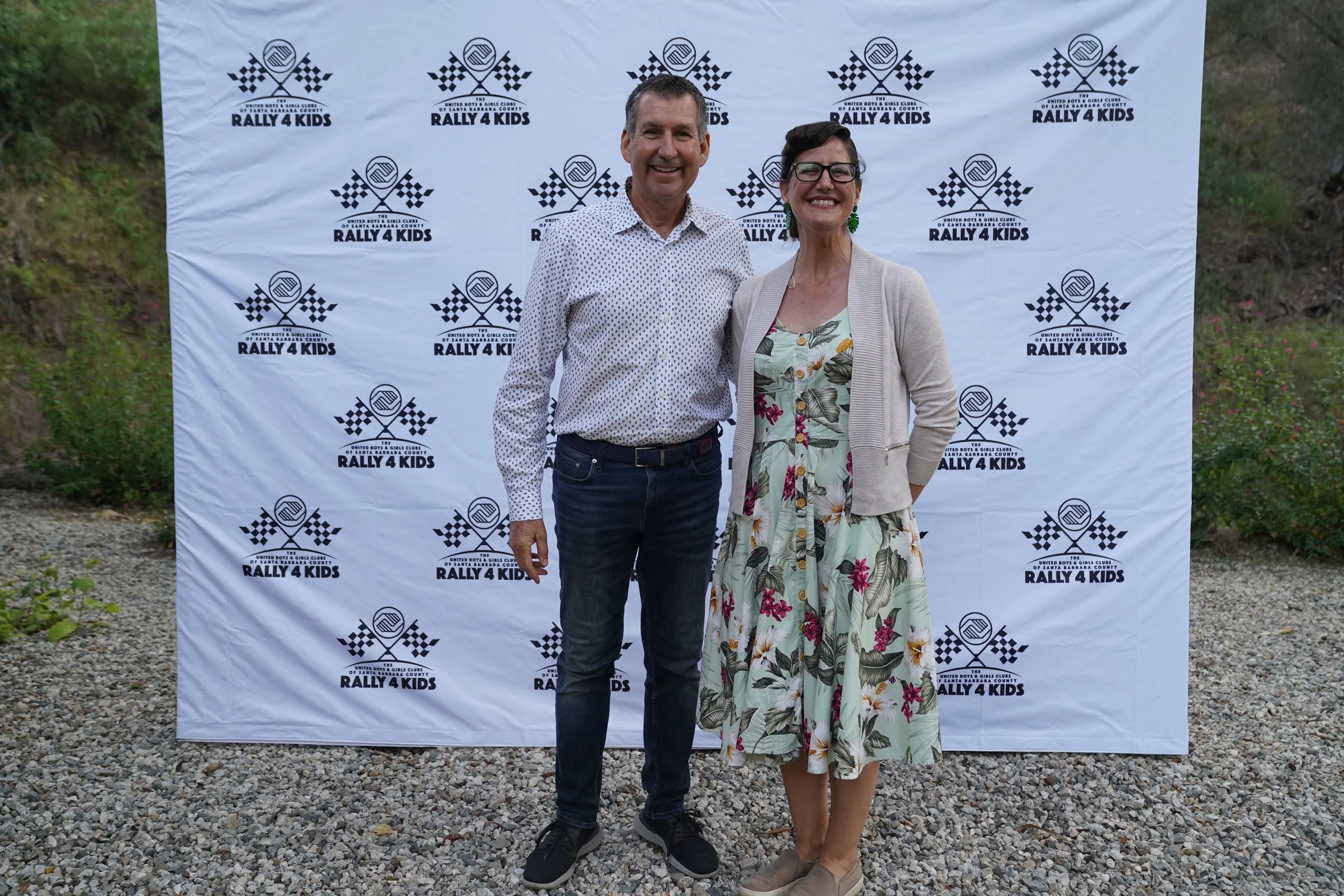 A man and a woman standing together outdoors in front of a backdrop with the Rally 4 Kids logo. They are smiling at the camera.