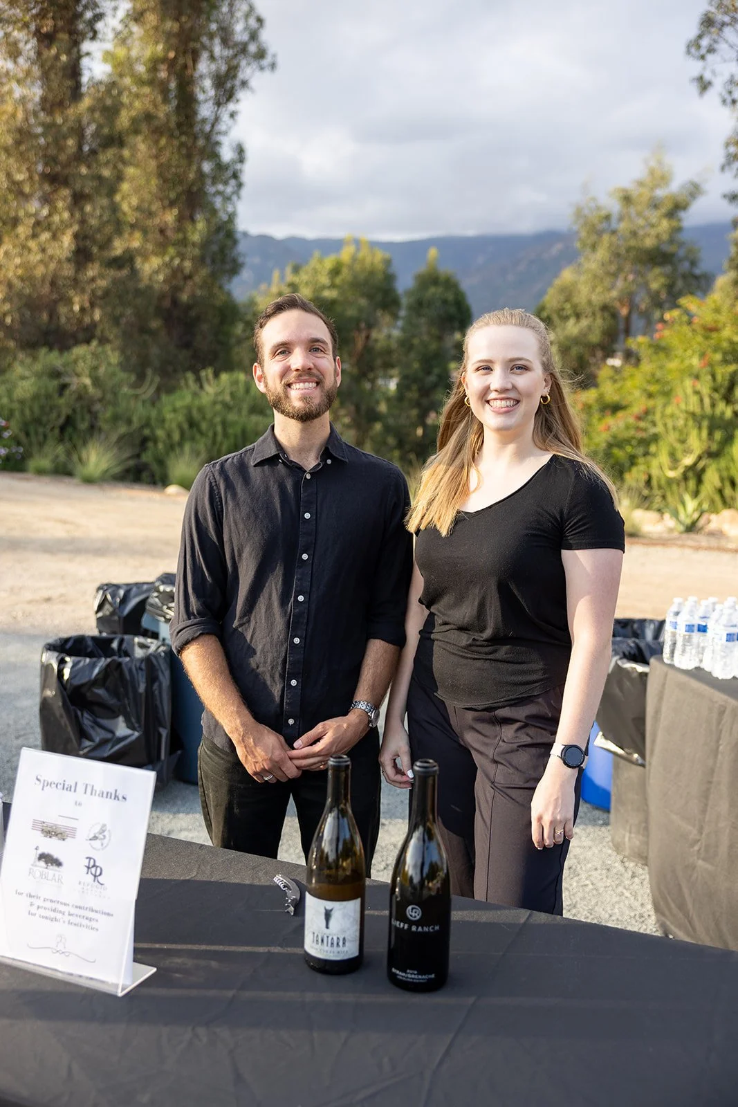 Two people, a man and a woman, standing behind a table with bottles of wine, at an outdoor event with trees, mountains, and cloudy sky in the background.
