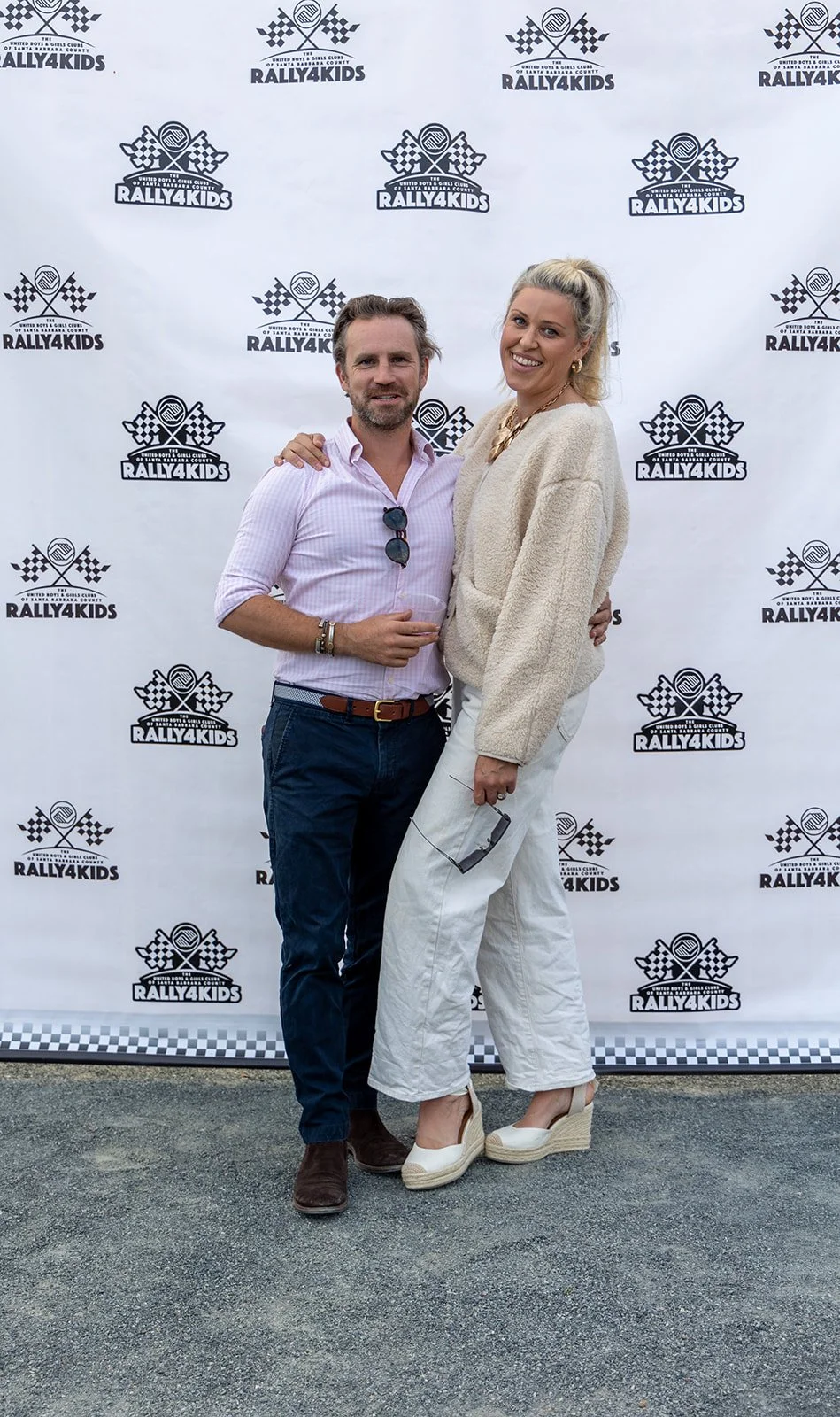 Two people standing in front of a Rally4Kids backdrop, smiling and posing for the photo. The man is wearing a light pink shirt, dark blue jeans, and brown shoes, while the woman is in a cream sweater, white pants, and white wedge shoes, holding sungl