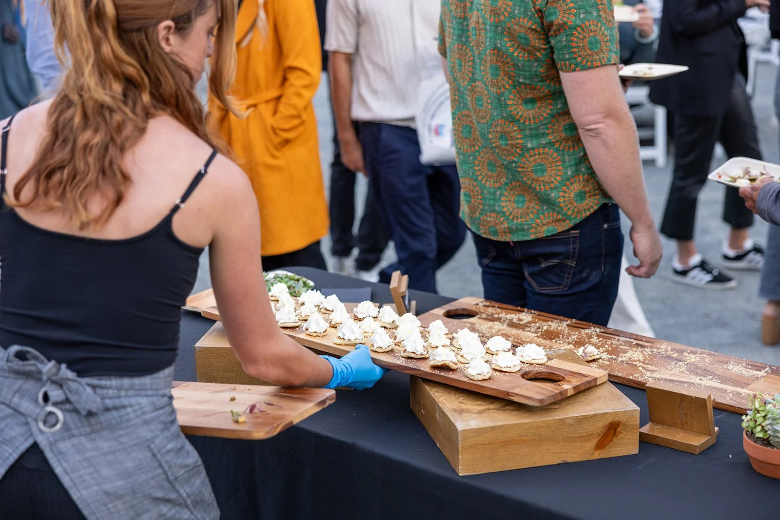 A woman in a black top serving food at an outdoor gathering, with plates of food on a wooden serving tray on a black table, surrounded by people in casual attire.