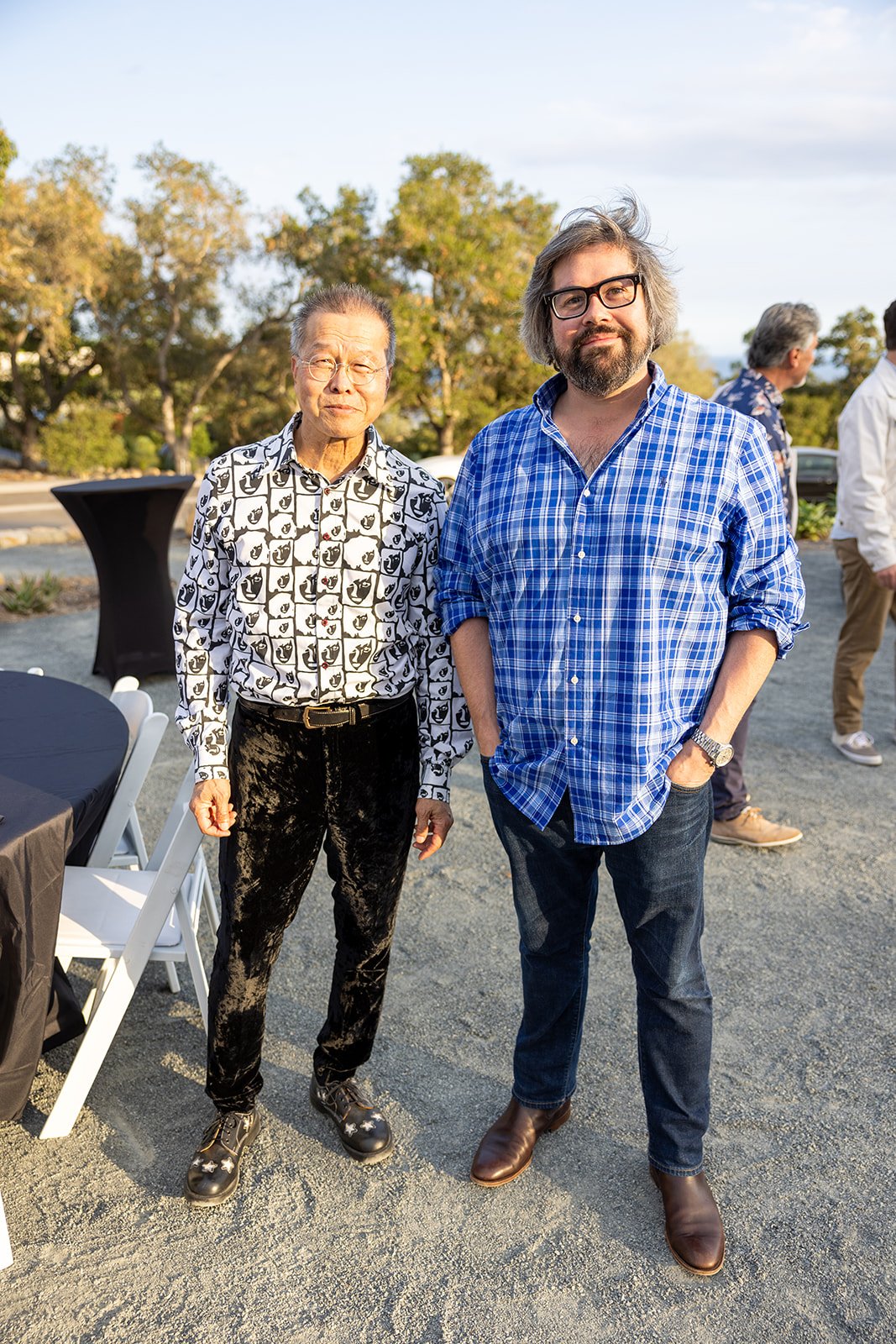 Two men standing outdoors at a social gathering. The man on the left is wearing glasses, a black and white patterned shirt, and black velvet pants. The man on the right has gray hair, a beard, and is dressed in a blue checkered shirt with rolled-up s