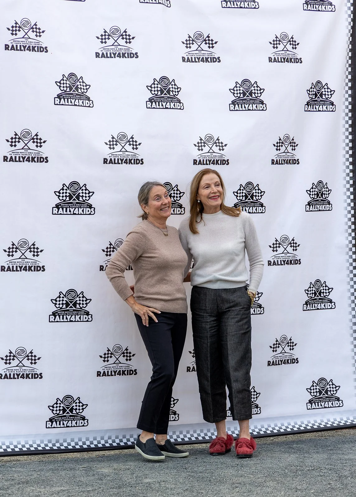 Two women standing in front of a Rally4Kids backdrop, smiling. One woman wears a beige sweater and black pants, the other wears a light gray sweater and checkered pants. They are posing with their arms around each other.