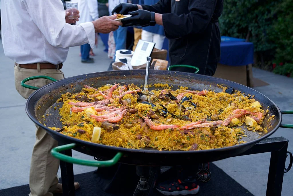 Large pan of cooked paella with rice, shrimp, and vegetables at outdoor event.