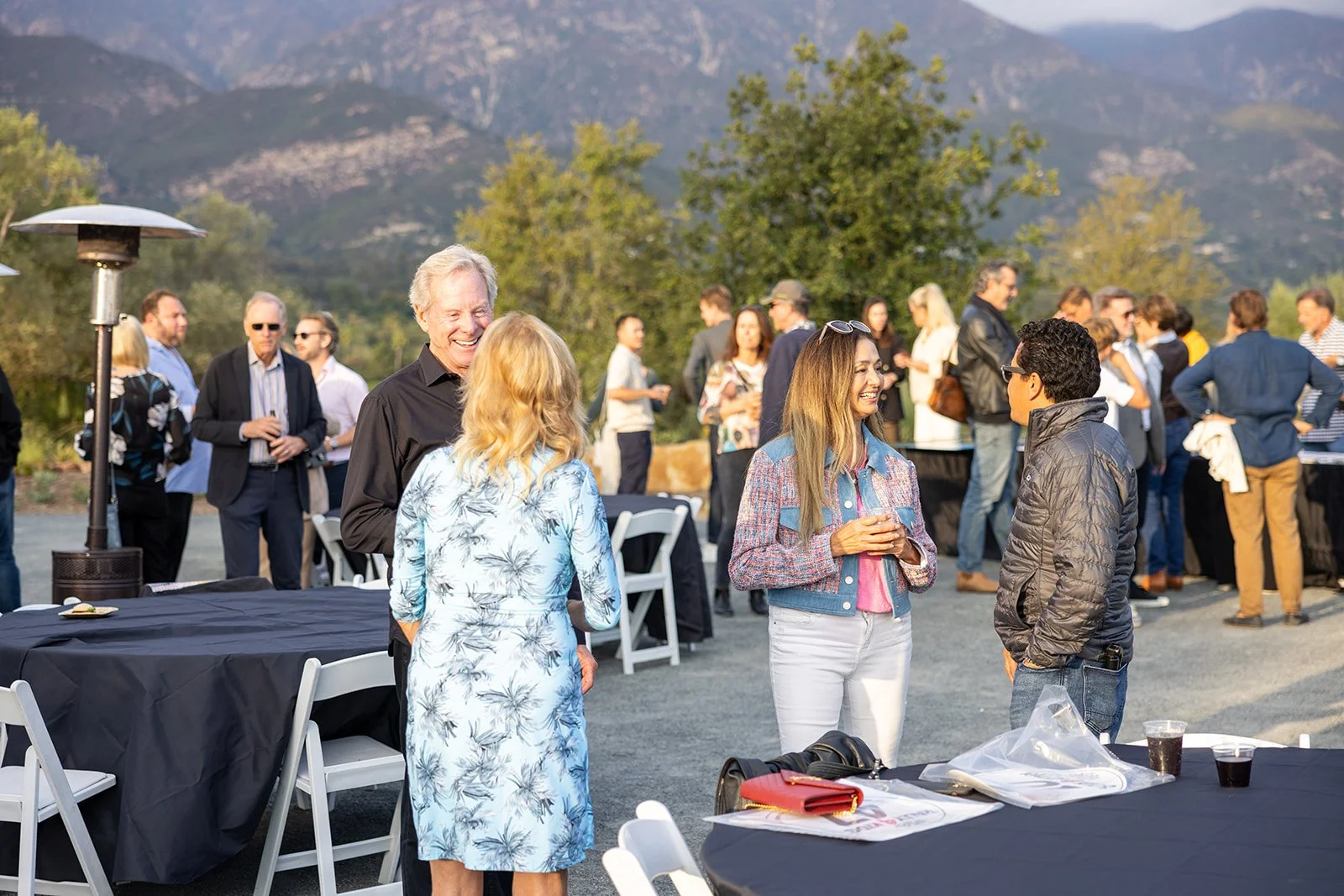 People socializing outdoors at a gathering with tables and mountain scenery in the background.