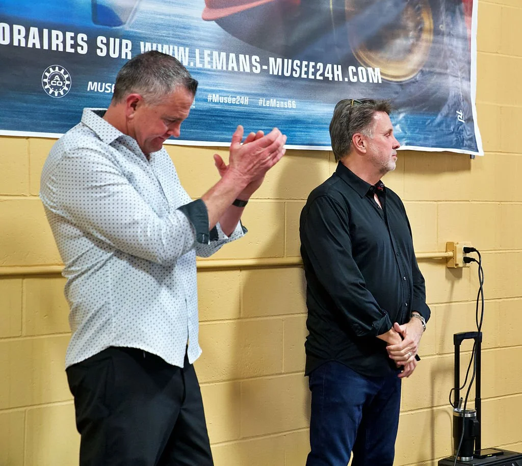 Two men standing against a yellow wall with a banner in the background, one clapping and the other looking down with hands clasped.