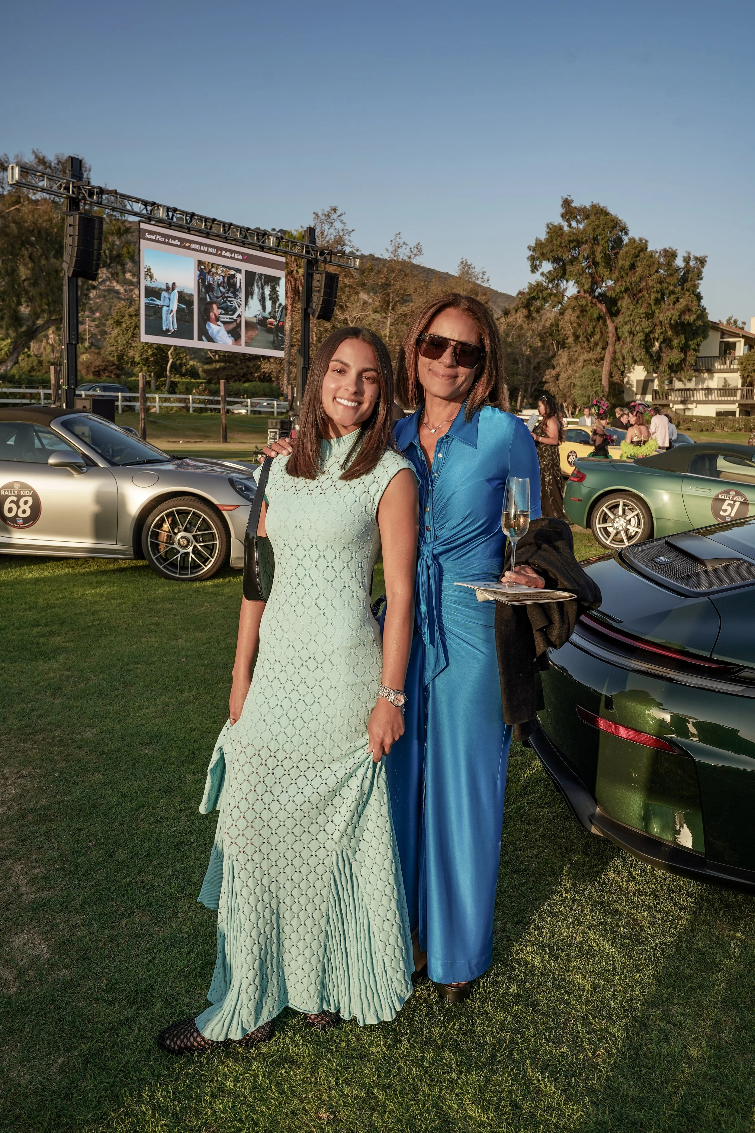 Two women standing on grass in front of luxury cars at an outdoor event, smiling for the camera. One woman is in a light-colored, patterned dress, while the other is in a blue outfit holding a glass of champagne.