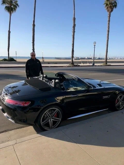 A man standing next to a black convertible sports car parked on the street near the ocean with palm trees and a pier in the background.