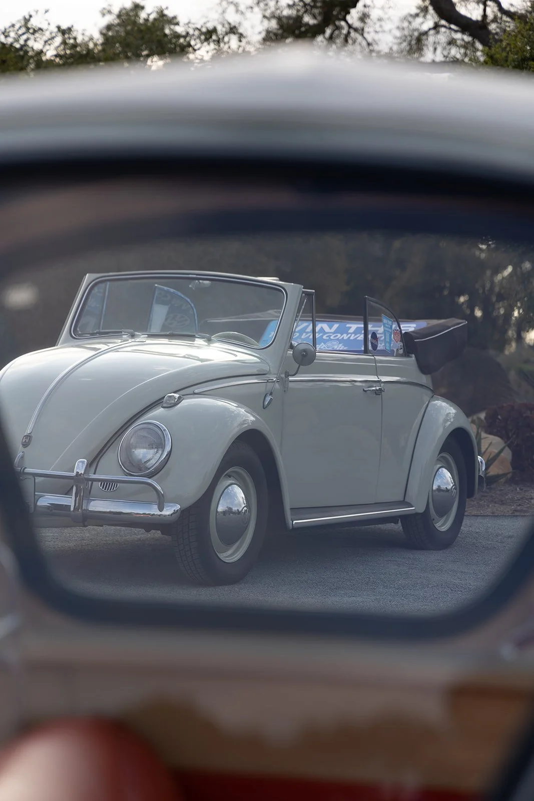 A vintage Volkswagen Beetle convertible viewed through a rearview mirror, with the car parked outdoors.