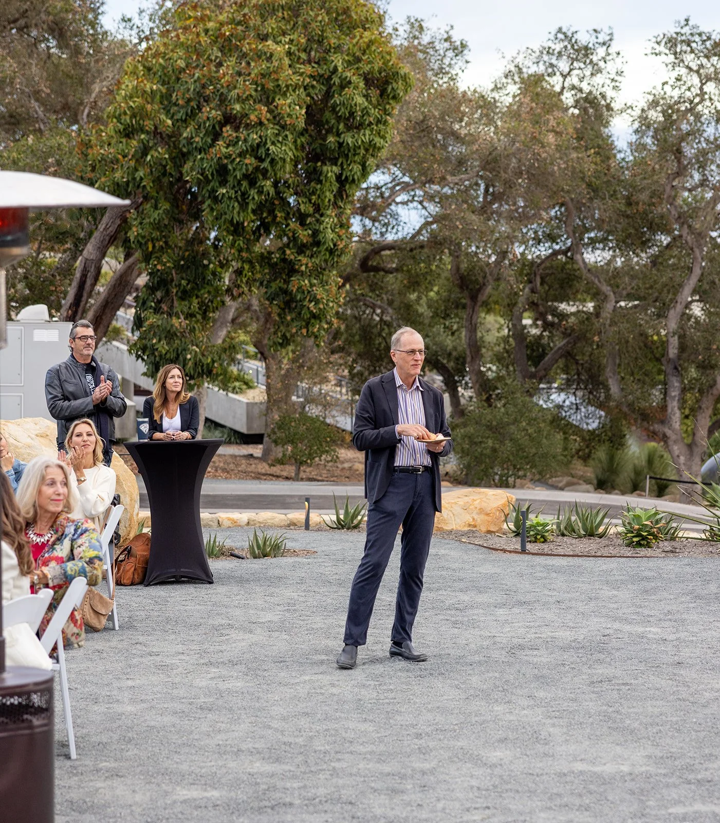 Man in a suit giving a speech at an outdoor event with seated and standing audience members, trees in the background.