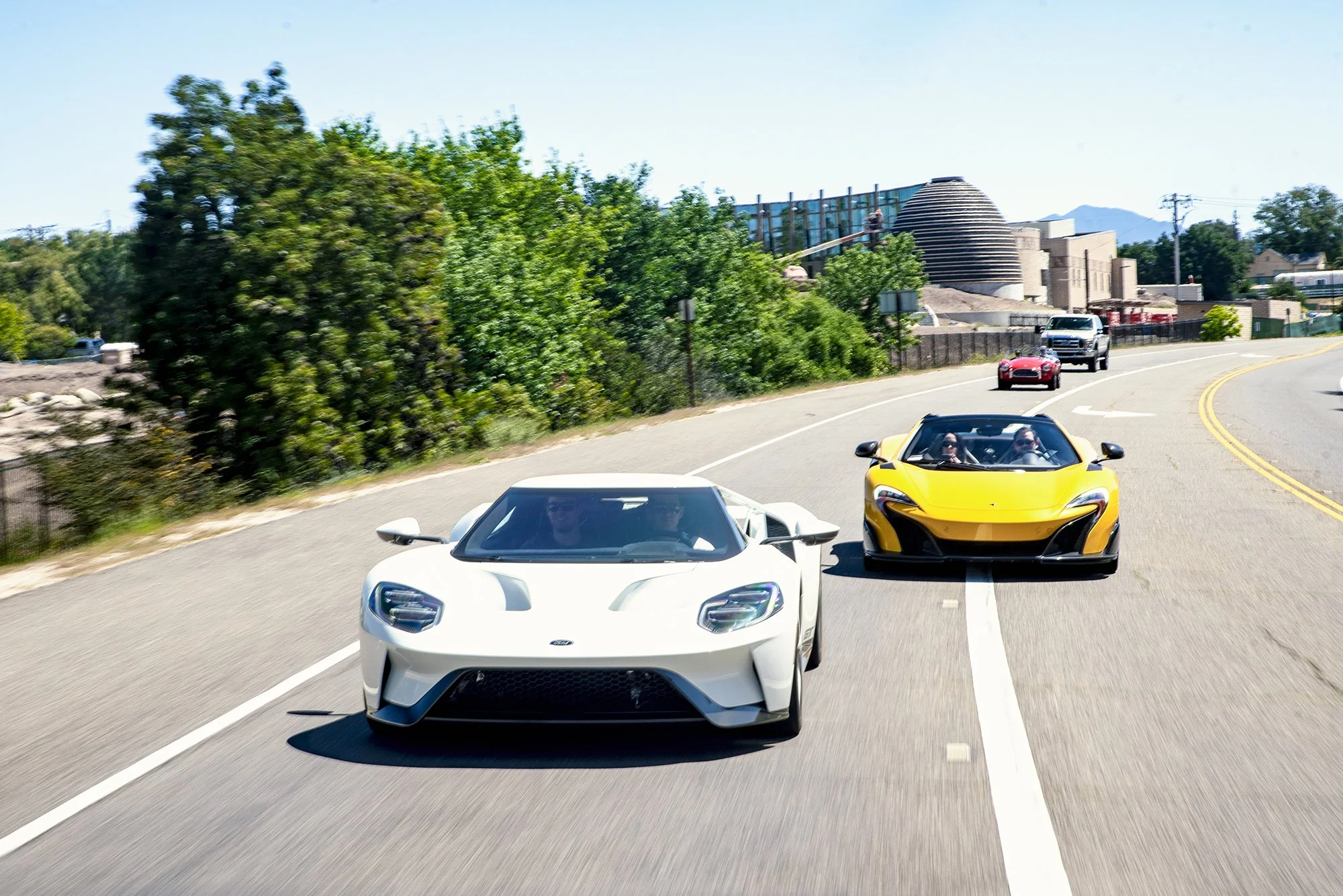 Two sports cars, one white and one yellow, driving on a curvy road with green trees on the side and a building and mountains in the background.