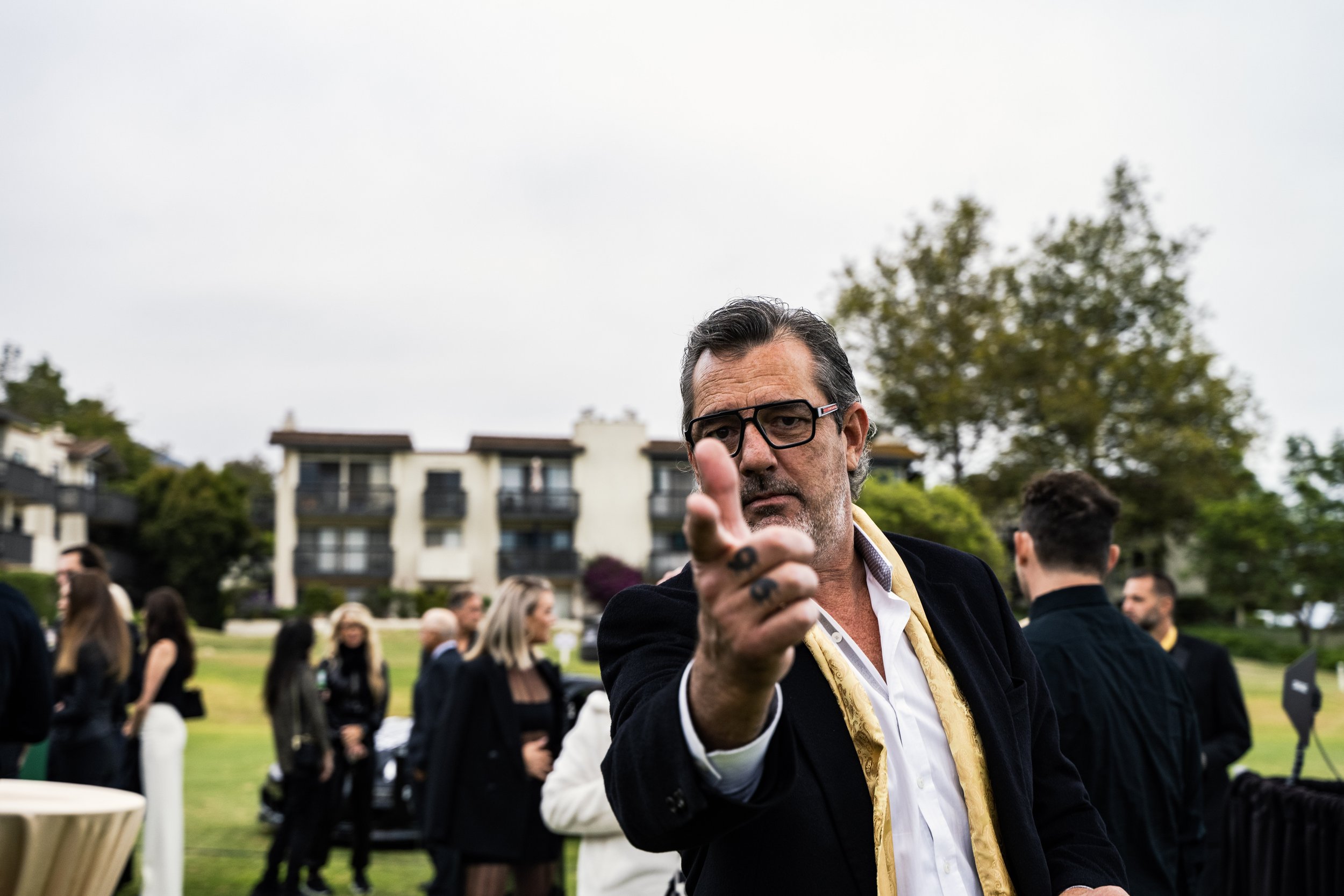 A man with glasses pointing towards the camera at an outdoor gathering with multiple people and a residential building in the background.