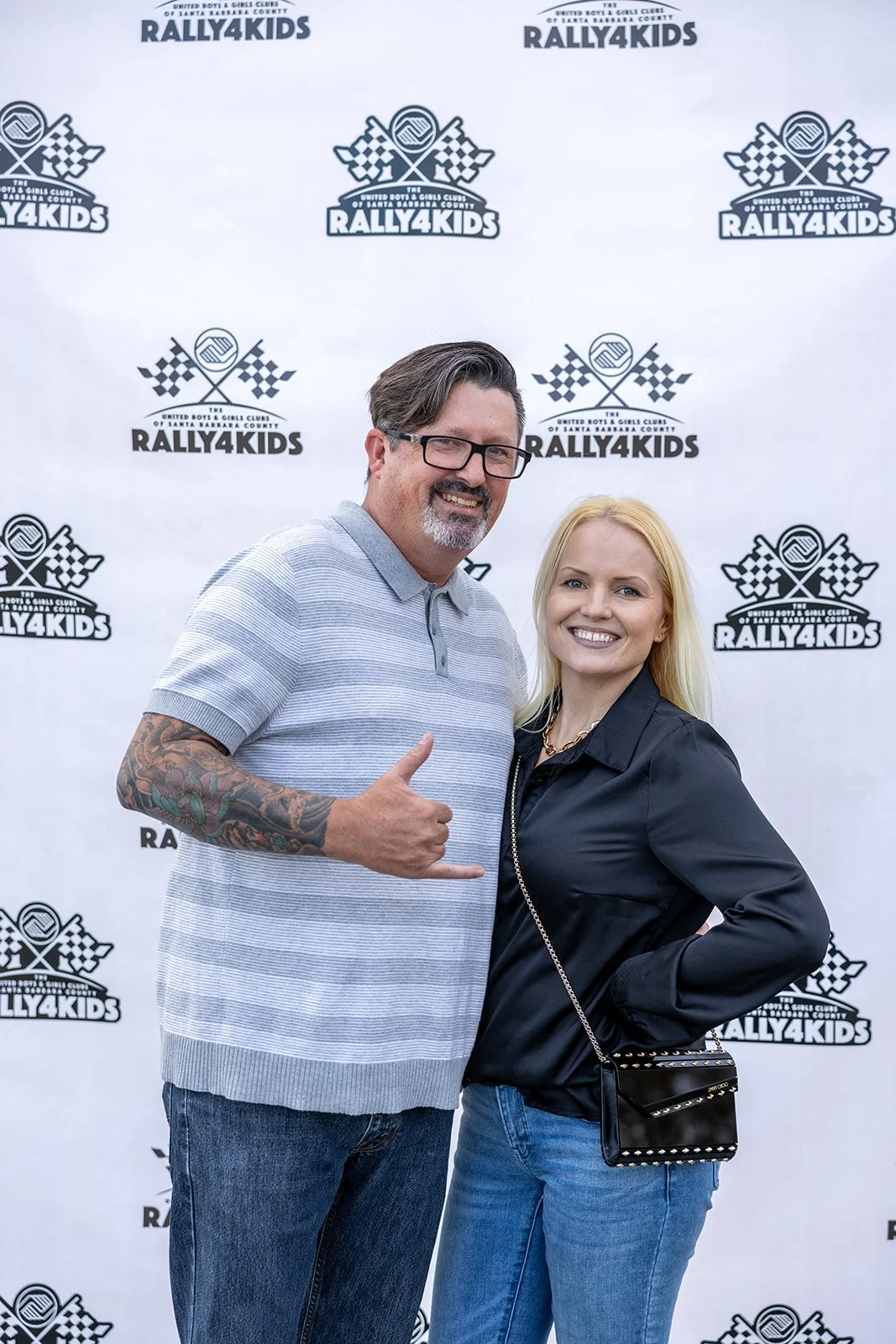 A man and woman standing in front of a Rally4Kids backdrop, smiling at the camera. The man has glasses, a beard, tattoos on his arm, and is giving a shaka sign. The woman has long blonde hair, is wearing a black jacket, and carrying a small black pur