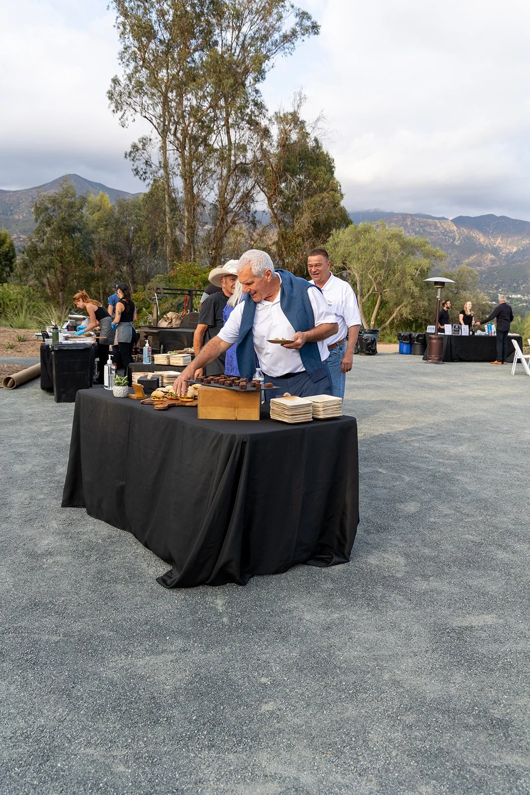 Guests at an outdoor event serving food, with mountains and trees in the background, during the daytime. One man is selecting food from a buffet table covered with black cloth, with plates and food items on it.